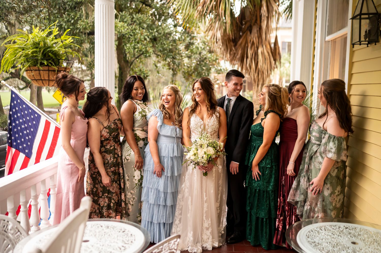 Wedding party in colorful dresses poses together on a yellow house porch with American flag and palm trees.