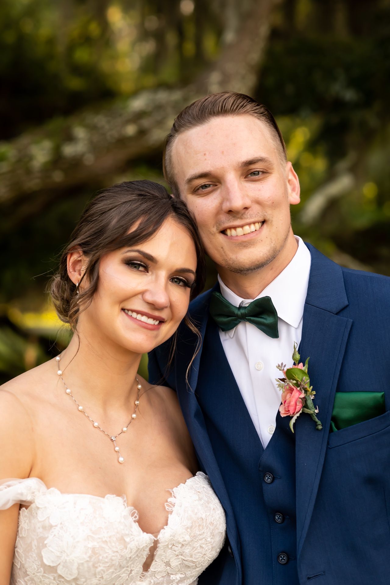 A close-up outdoor portrait of a bride in a lace dress and groom in a navy suit with bow tie at their wedding.