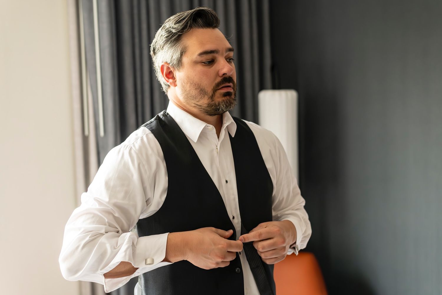 Businessman in white dress shirt and black vest buttoning up his formal attire in a room.