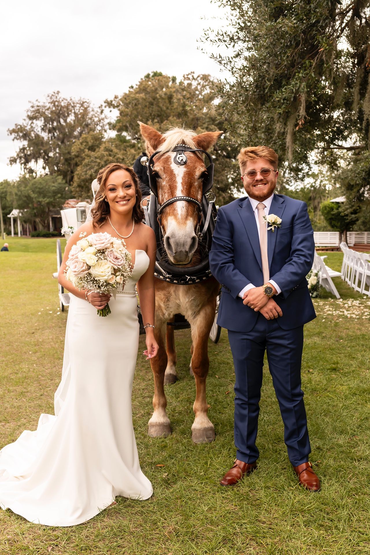 A couple poses in wedding attire with their large brown draft horse on a grassy lawn surrounded by trees.
