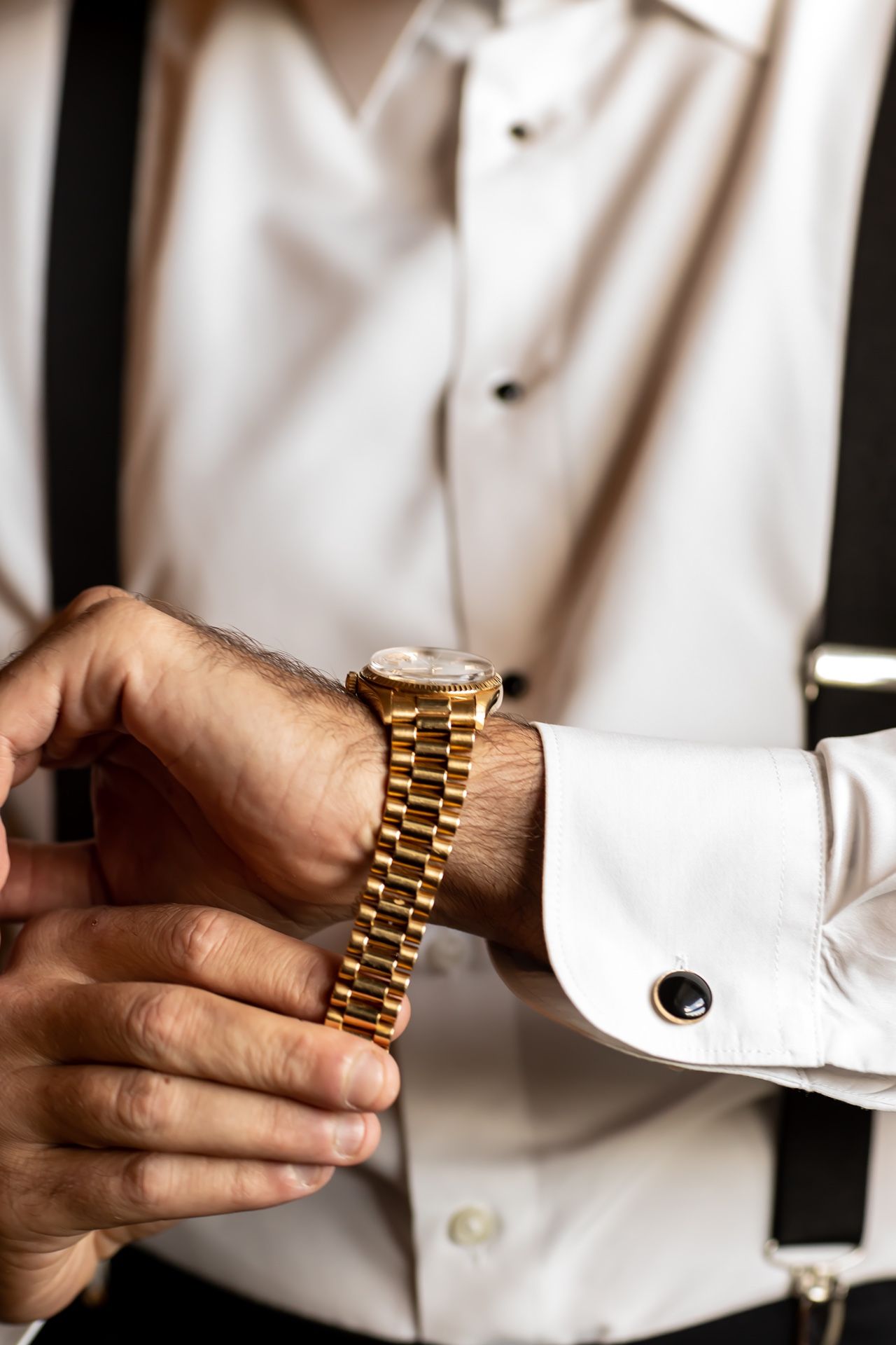 Close up shot of hands adjusting a gold watch with a formal white dress shirt and black suspenders visible.