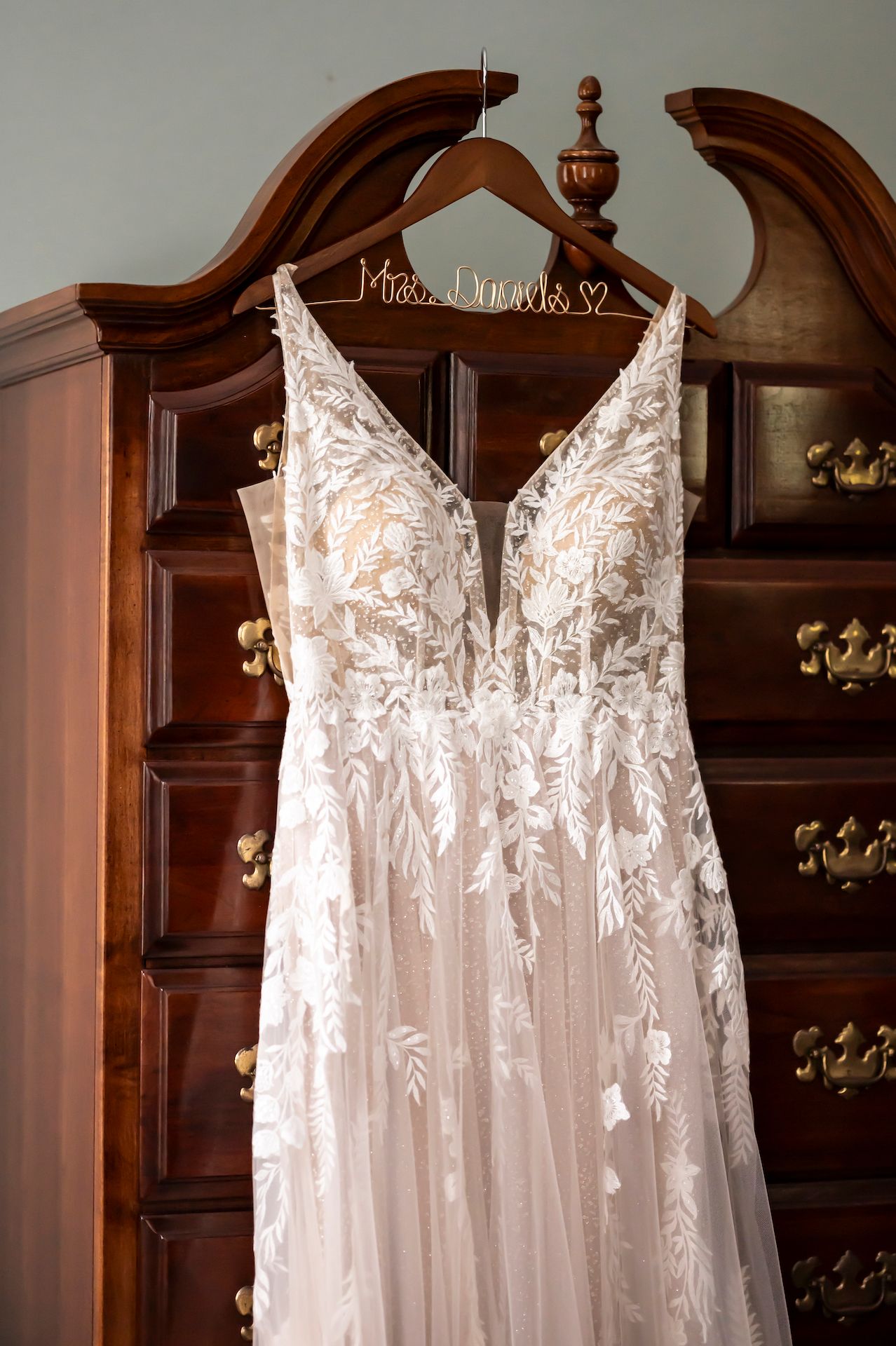 Elegant lace wedding dress with plunging neckline hangs on personalized wooden hanger against ornate wooden wardrobe.