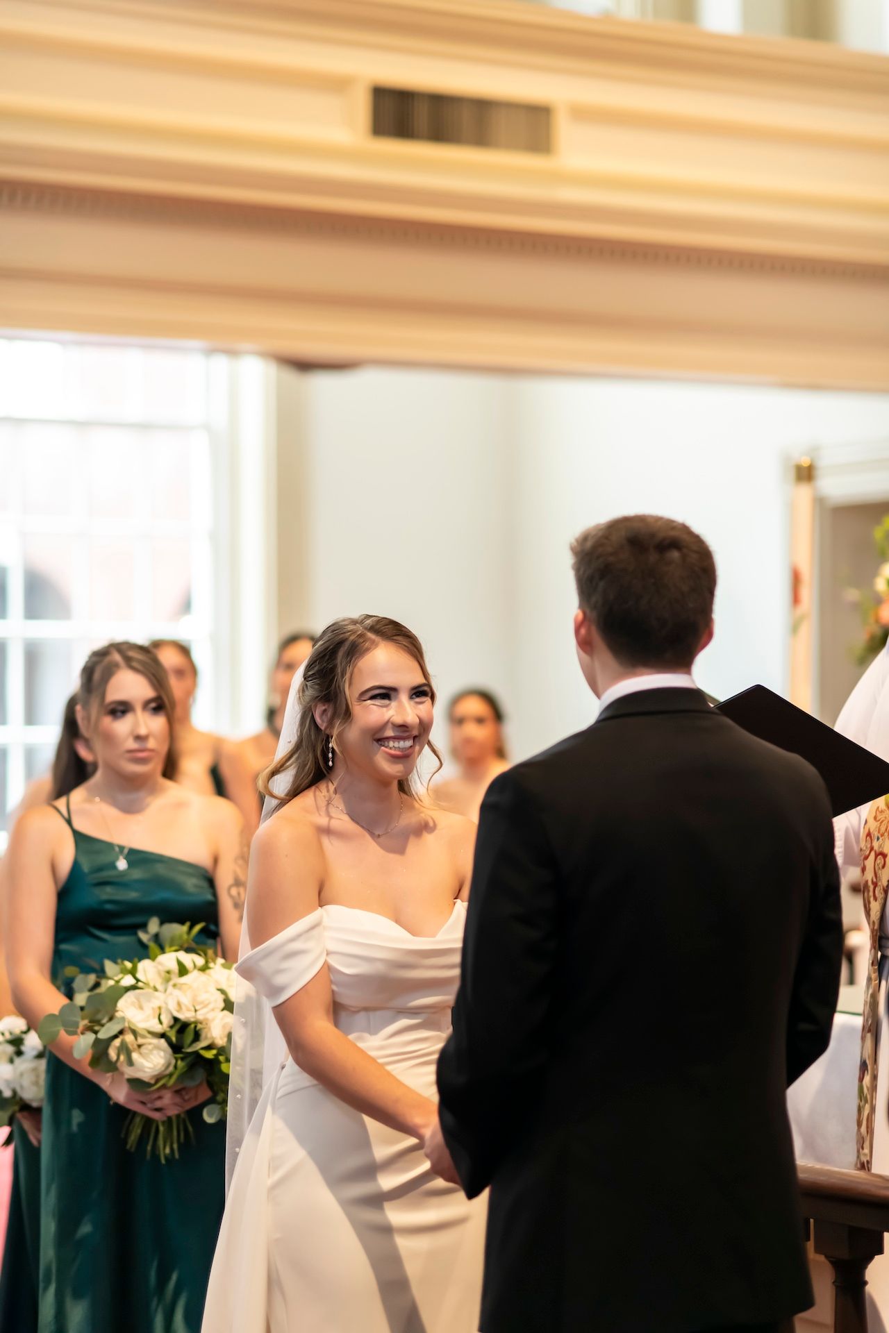 Bride and groom exchange vows at altar during indoor wedding ceremony with bridesmaid in emerald dress.