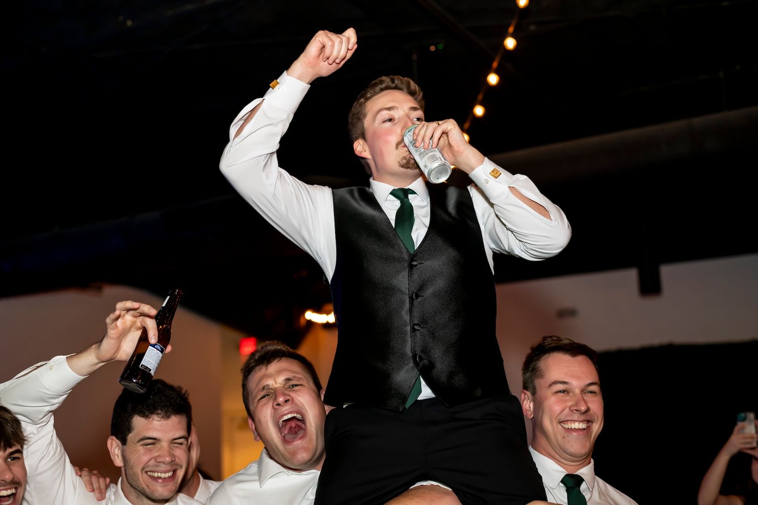 Energetic performer stands on chair singing into microphone while celebration crowd cheers at indoor party venue.