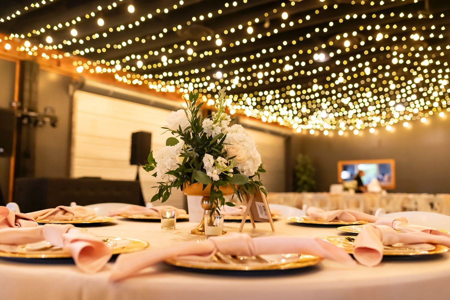 Elegant wedding reception table setting with string lights overhead and floral centerpiece on table with pink napkins.