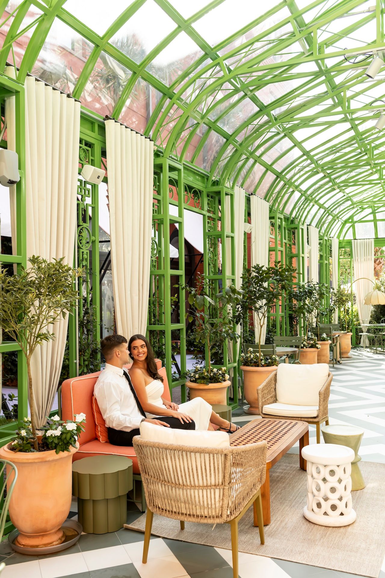 Two people relax in a luxurious conservatory with green metal framing, tall white curtains and potted plants.