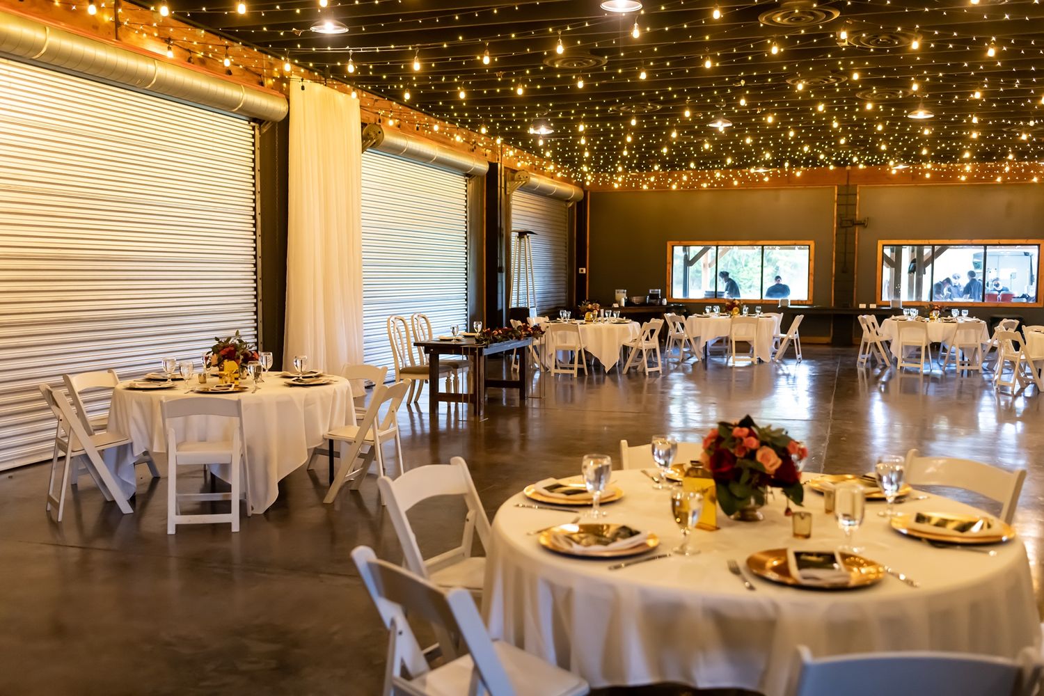 Elegant wedding reception venue with round tables, string lights, and white chairs in an industrial-style space.