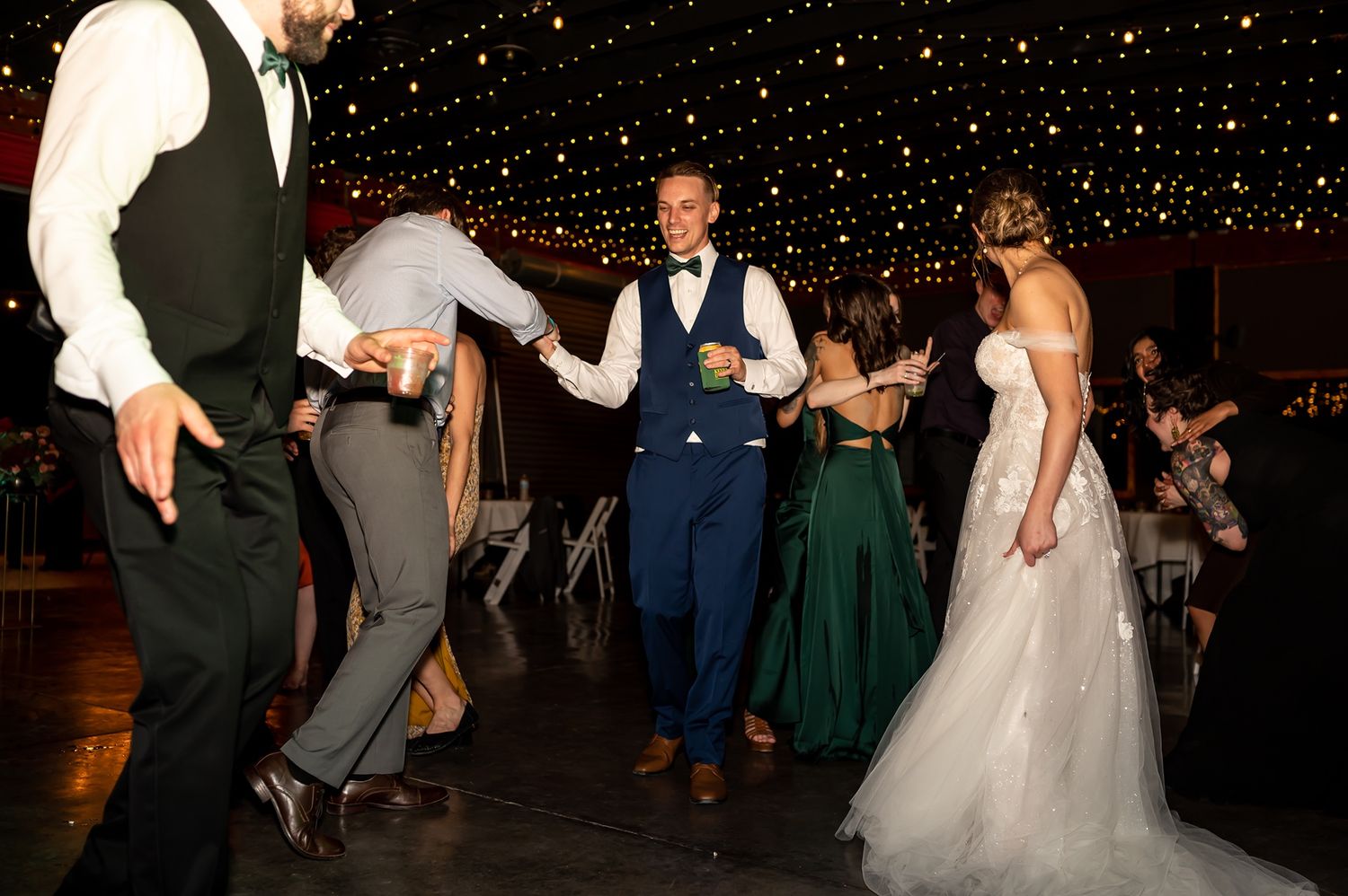Wedding party guests dance together on a dark dance floor with twinkling lights during a fun reception celebration.