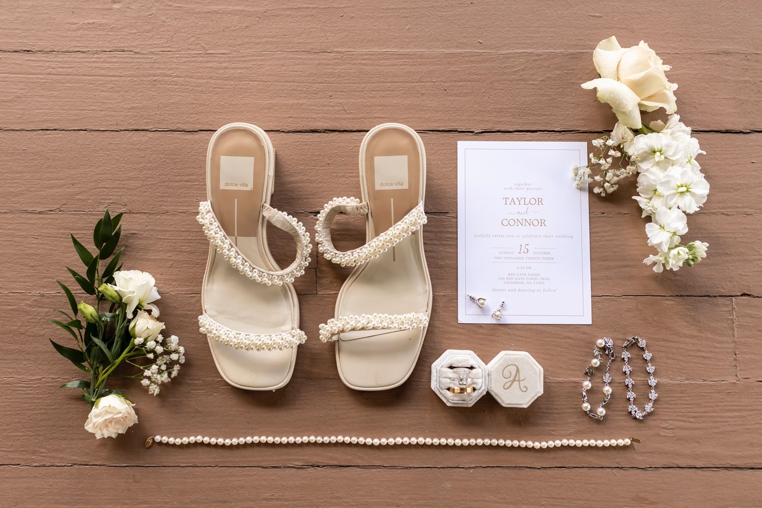 Wedding accessories including white sandals, floral bouquets, invitation, pearls, and jewelry laid on brown wooden surface.