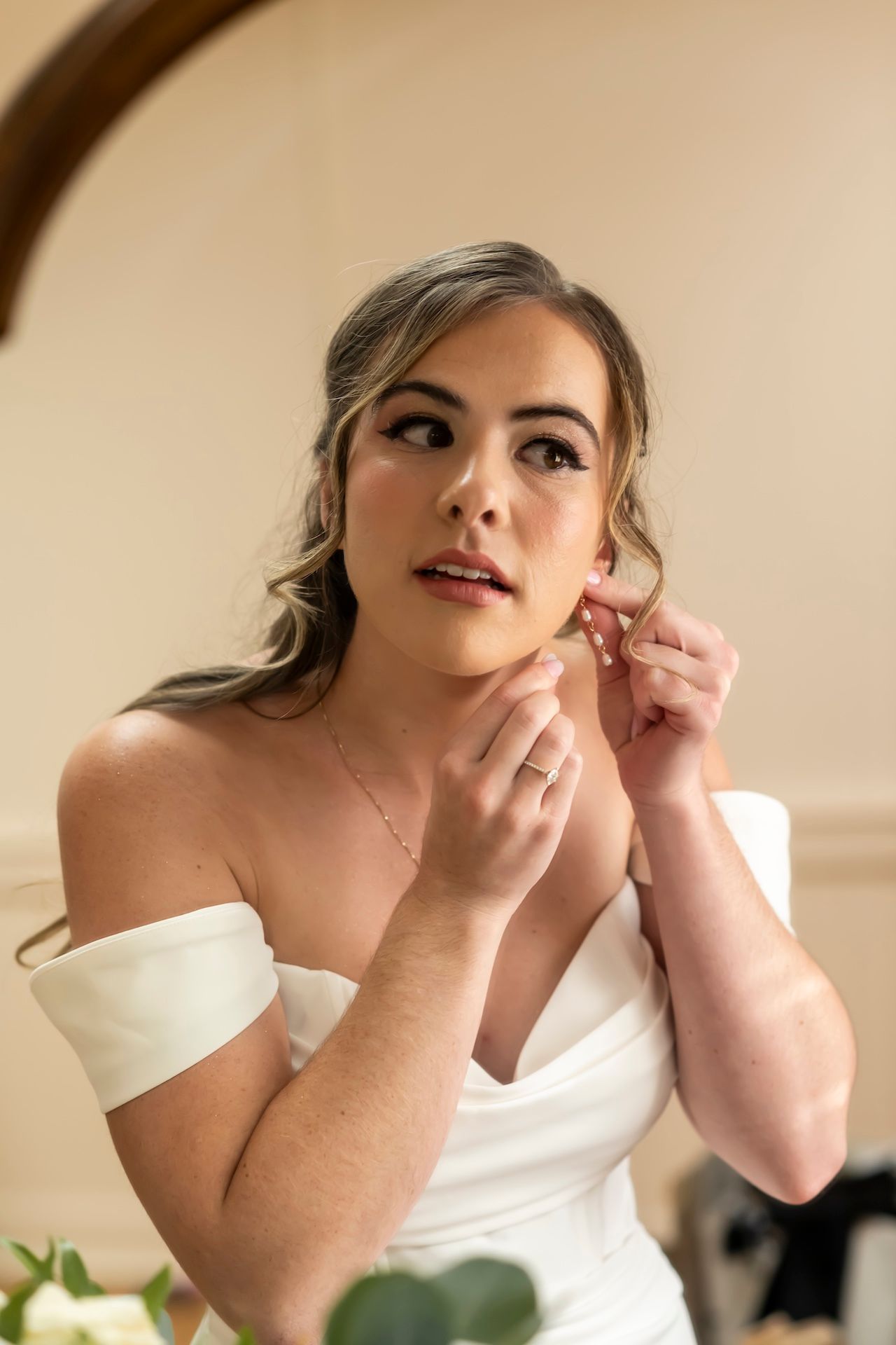 Someone in an off-shoulder white wedding dress puts on earrings while getting ready, with soft lighting and greenery visible.