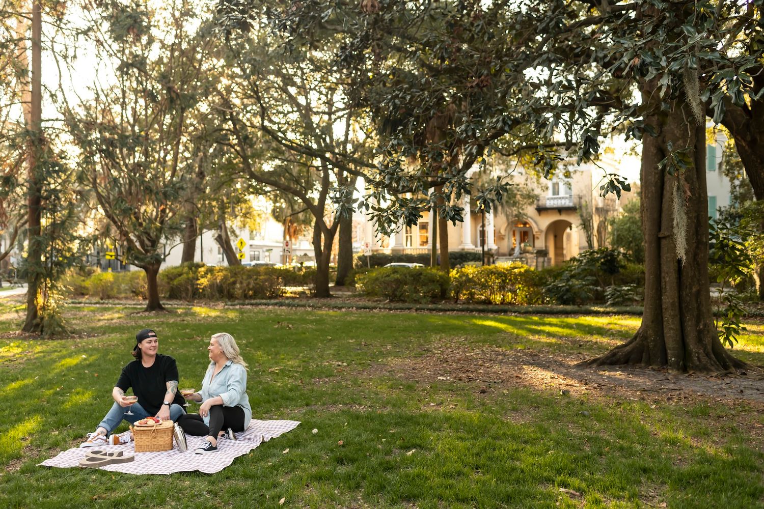 Two people enjoy a romantic picnic on a blanket in a sunlit park with Spanish moss draped trees and historic buildings.
