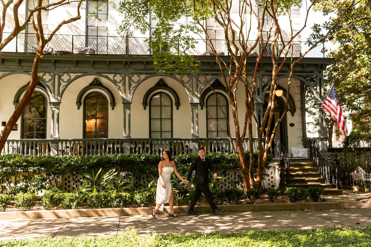 Historic white antebellum mansion with arched windows and wraparound porch in lush garden setting with American flag.