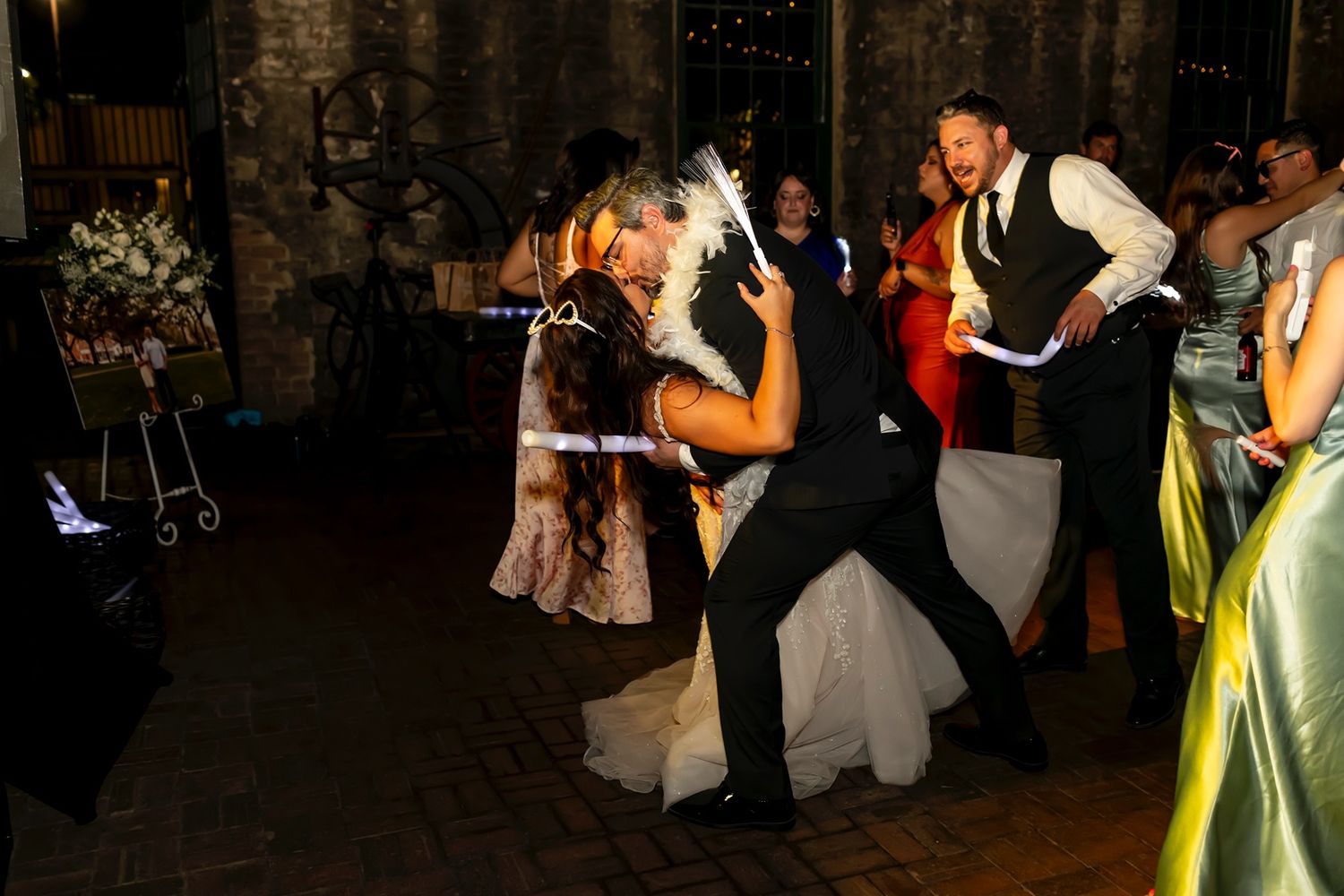 A couple dancing energetically at a wedding reception inside a rustic venue with dim lighting and other guests in the background.