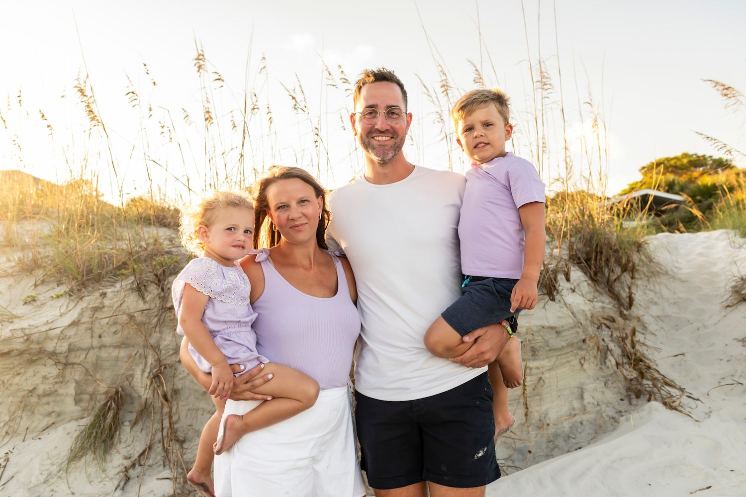 A loving family poses together on a sandy beach dune at sunset with tall grass swaying in the background.