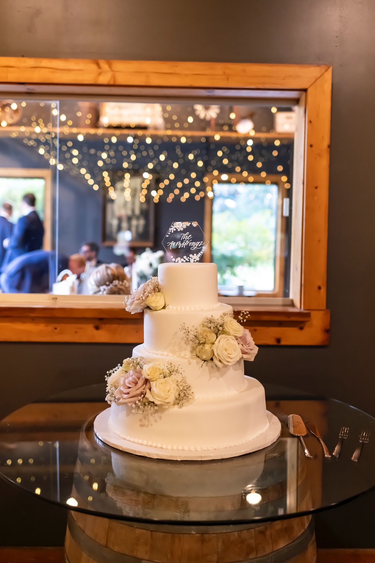 Elegant three-tiered white wedding cake with blush floral decorations displayed on glass table with twinkling lights in background.