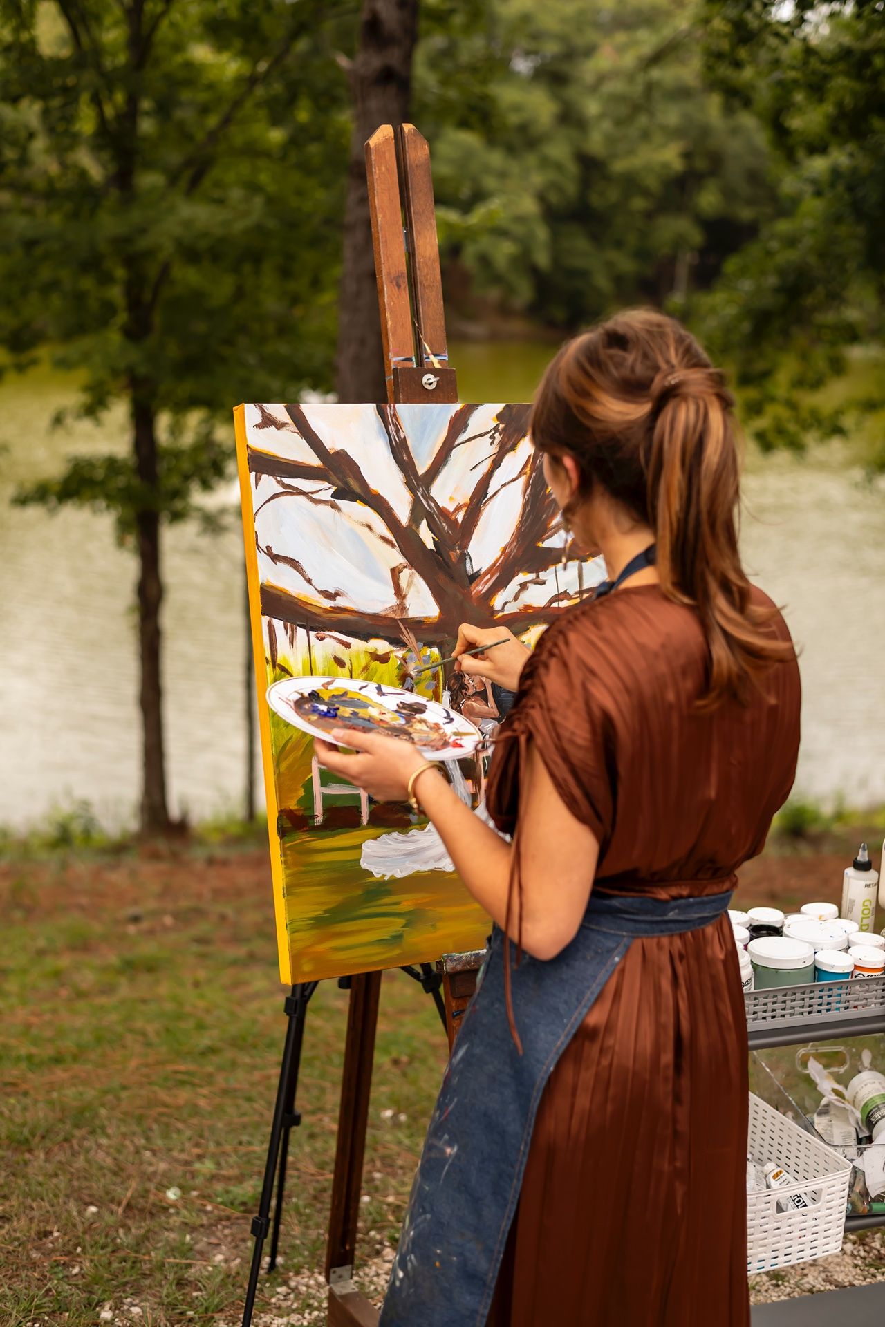 Artist in brown dress and denim apron paints a tree scene on canvas at outdoor easel with palette in hand.