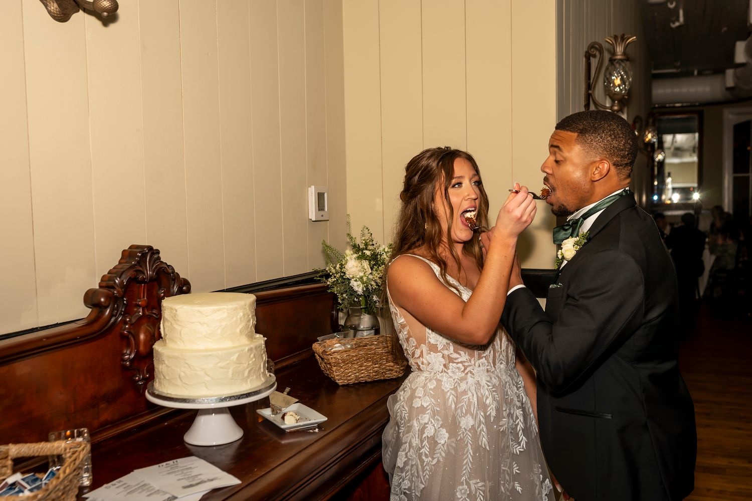 A newlywed couple share a playful moment feeding each other wedding cake at their reception.