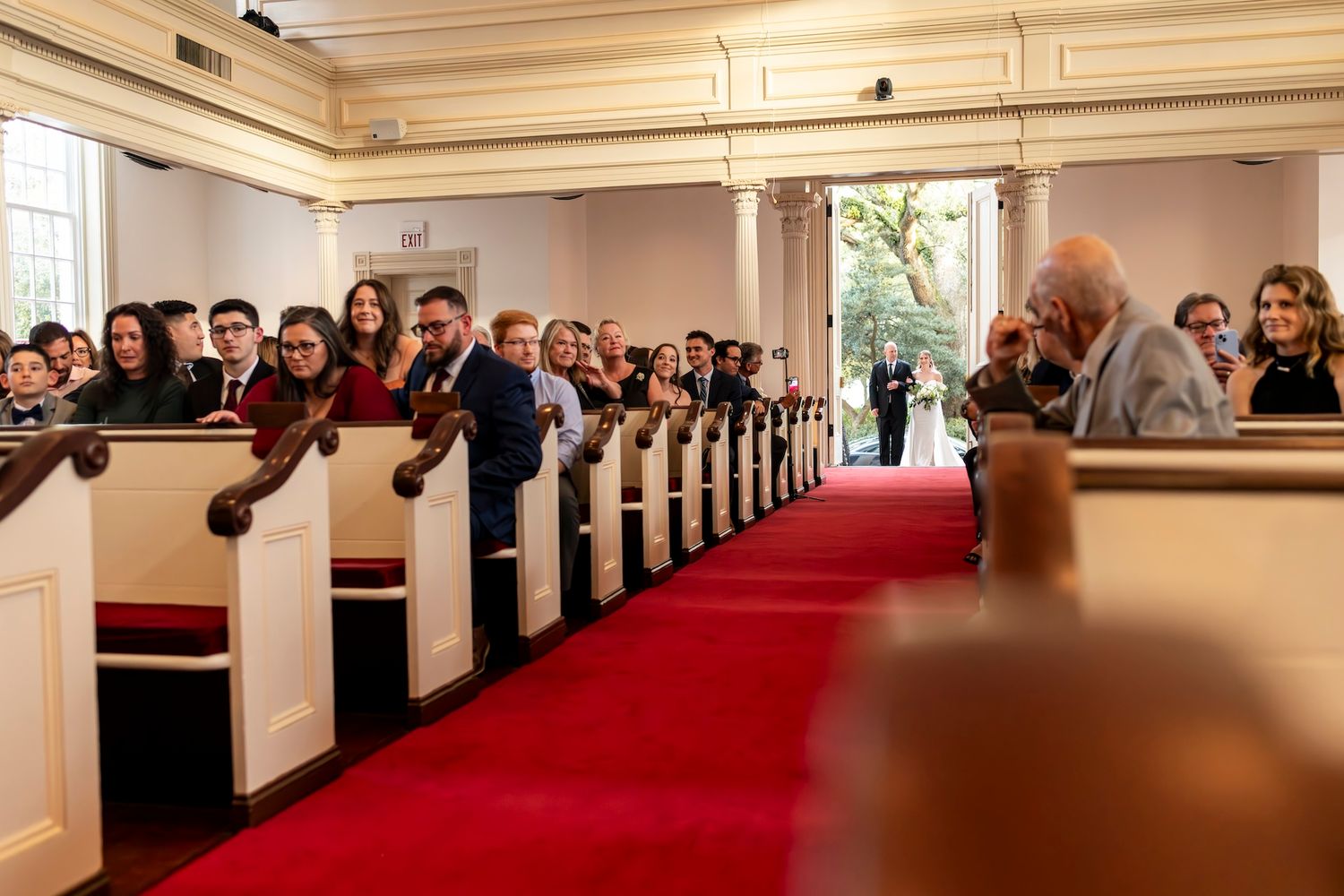 Wedding guests seated in white wooden pews with red carpeted aisle in traditional church sanctuary.