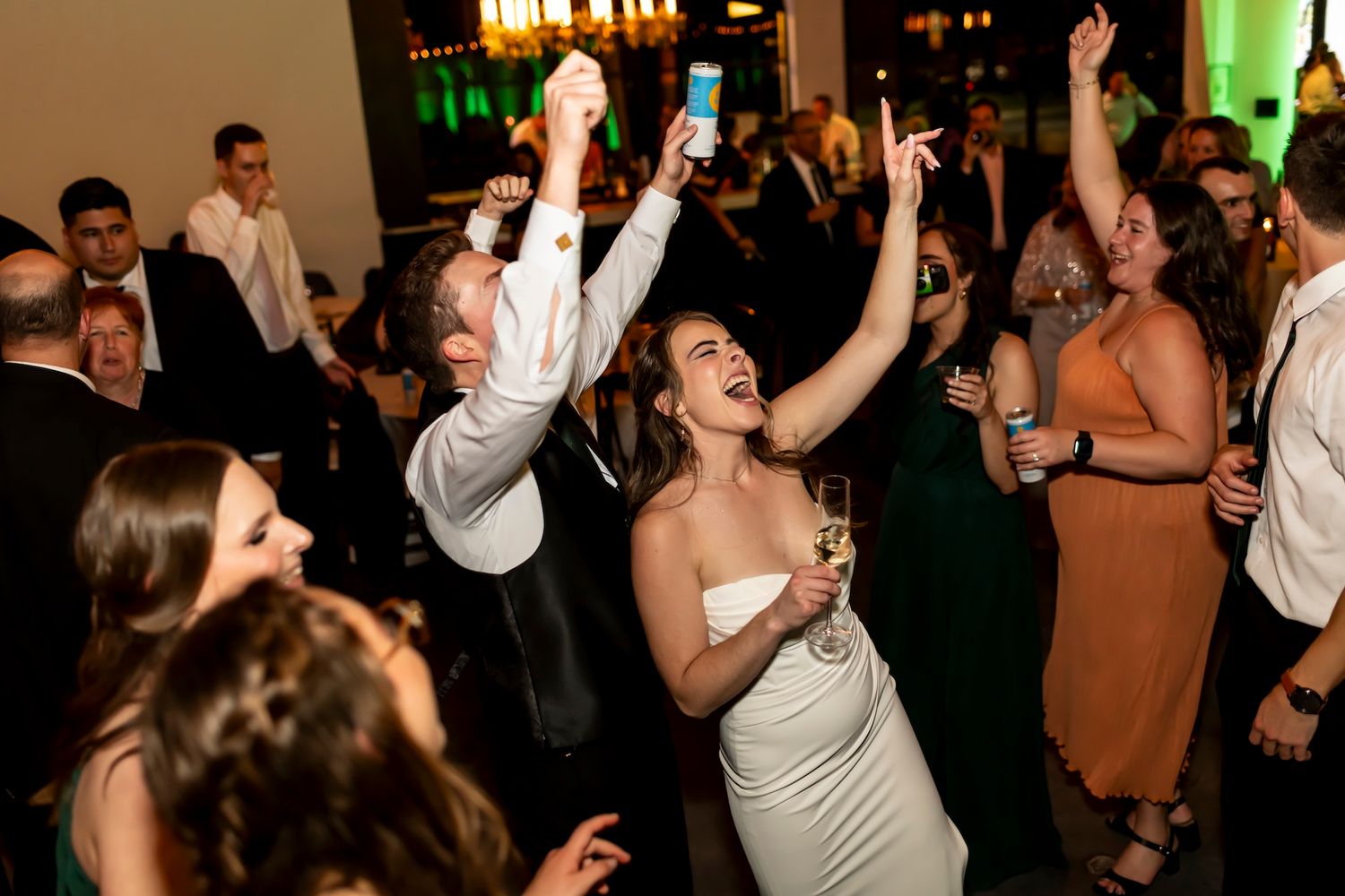 Wedding guests dance enthusiastically on the dance floor during a lively reception celebration with colorful lighting.