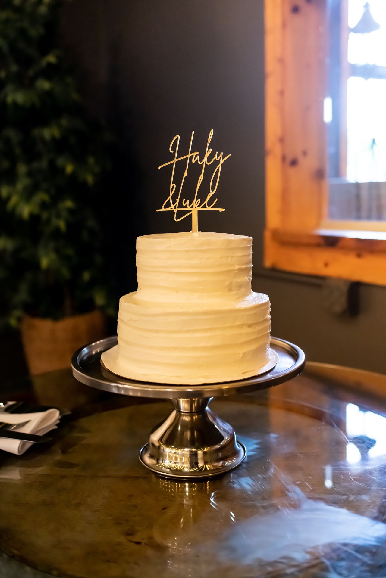 Two-tiered white wedding cake with gold 'Holy Light' topper displayed on silver stand.