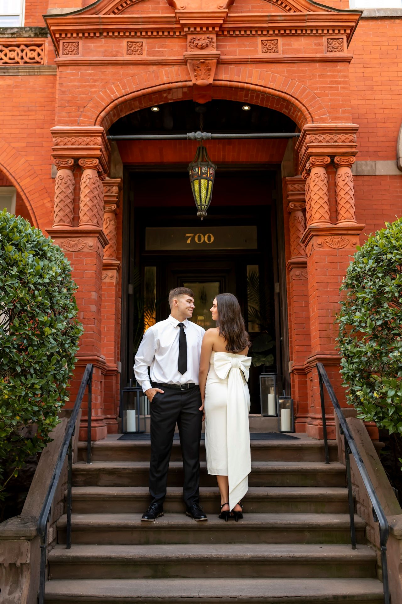A couple stands on ornate brick steps in front of a historic red building entrance with decorative archway and lantern.