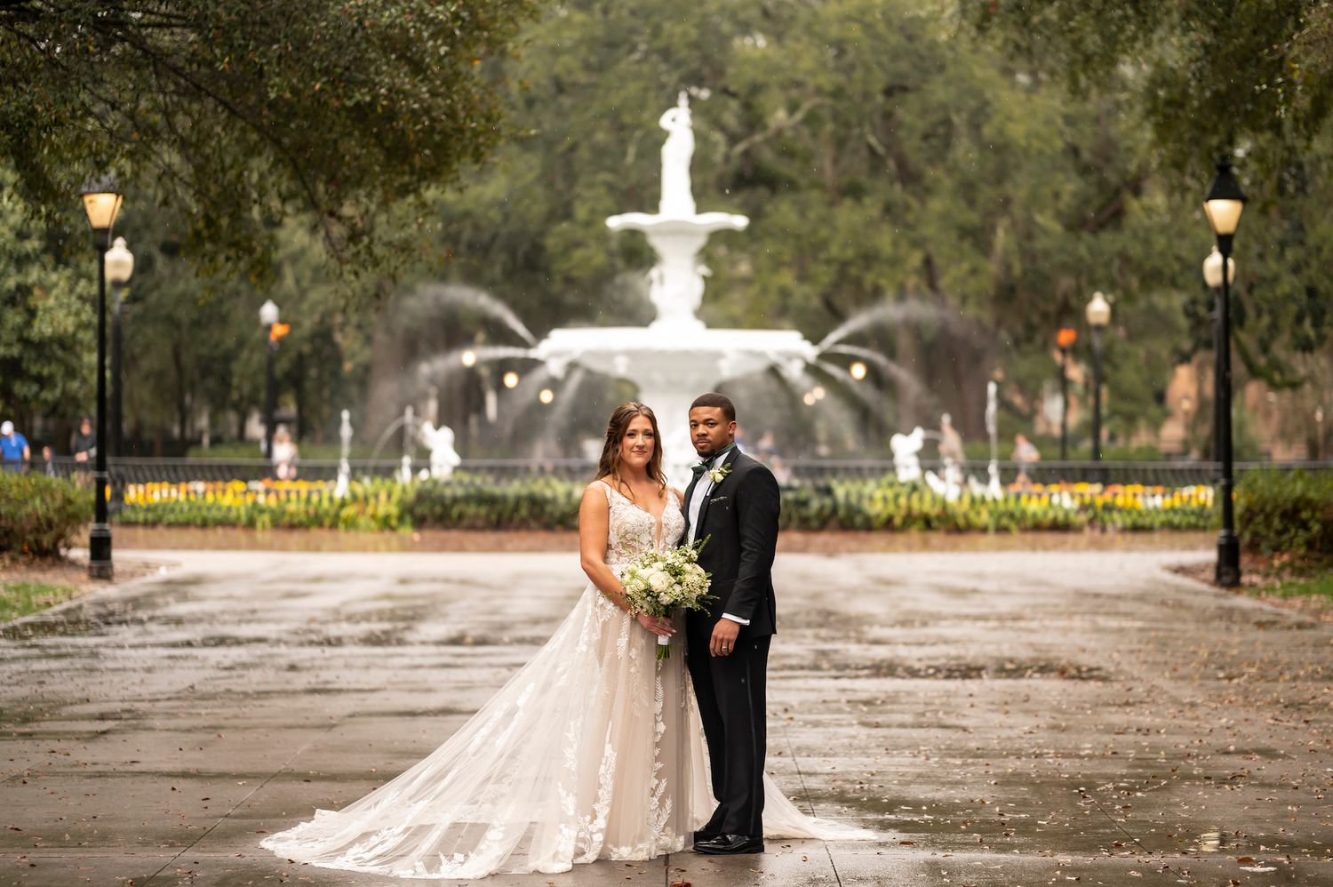 A couple in wedding attire stands on a pathway in front of the iconic Forsyth Park fountain in Savannah, Georgia.