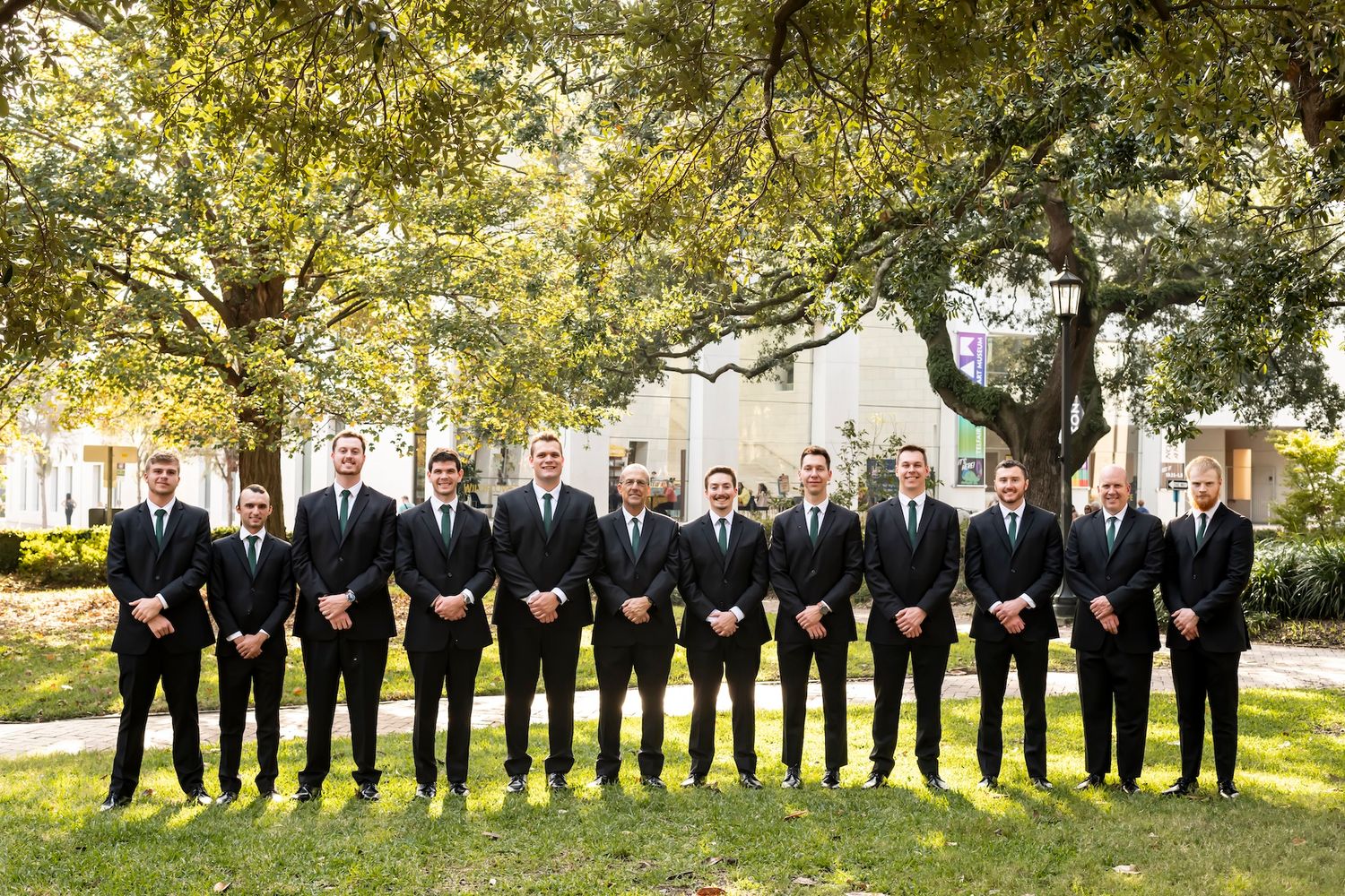 A line of men in black suits and ties stand together on a sunny lawn with trees in the background.