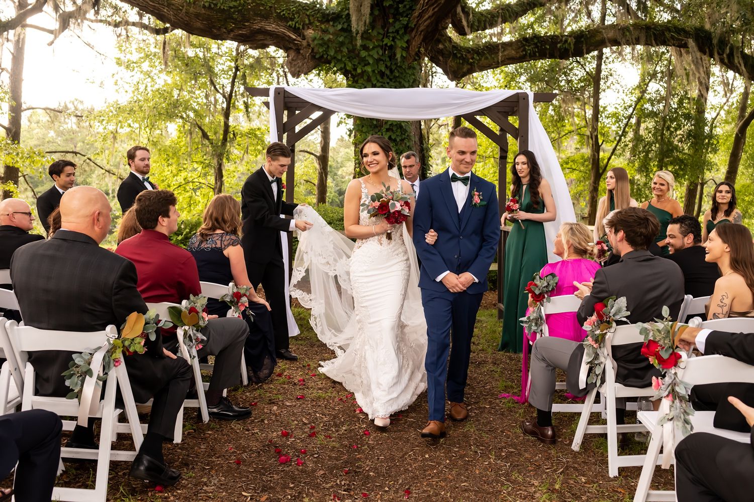 Couple walks down aisle at outdoor garden wedding ceremony under white chiffon arch with folding chairs and guests.