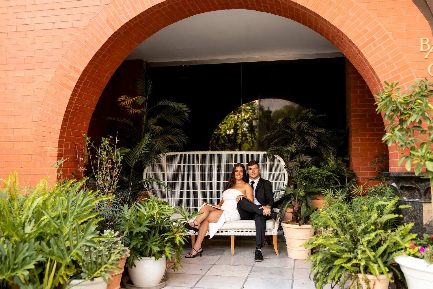 A couple sits together on a white peacock chair under a terracotta archway surrounded by lush green plants and flowers.