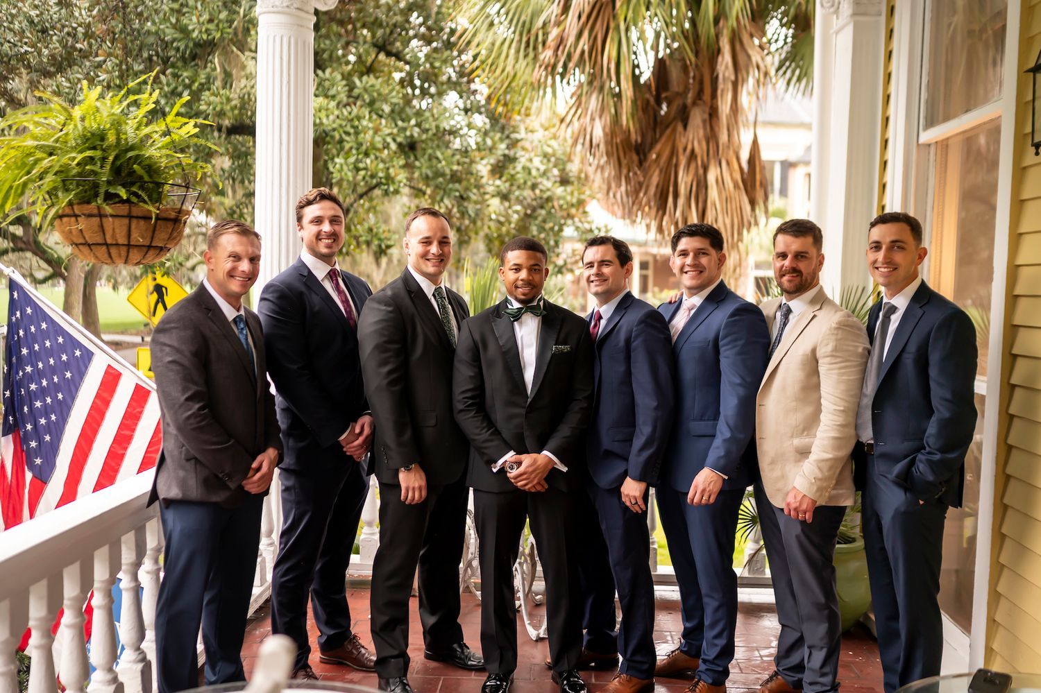A group of well-dressed men in suits stand together on a porch with an American flag displayed.