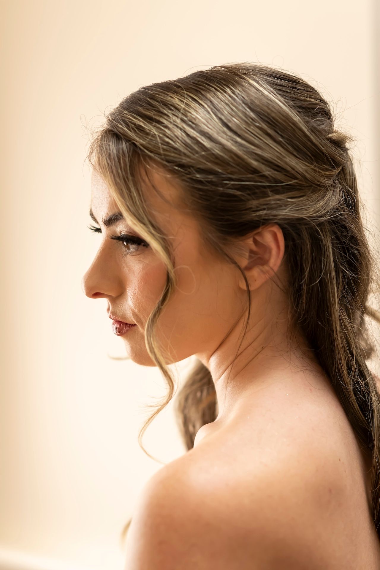 Profile view of a woman with flowing brown hair in soft lighting with bare shoulders against a warm beige background.