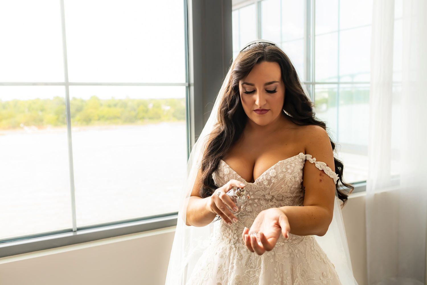 A bride in an elegant off-shoulder lace wedding gown stands by a window in natural light.