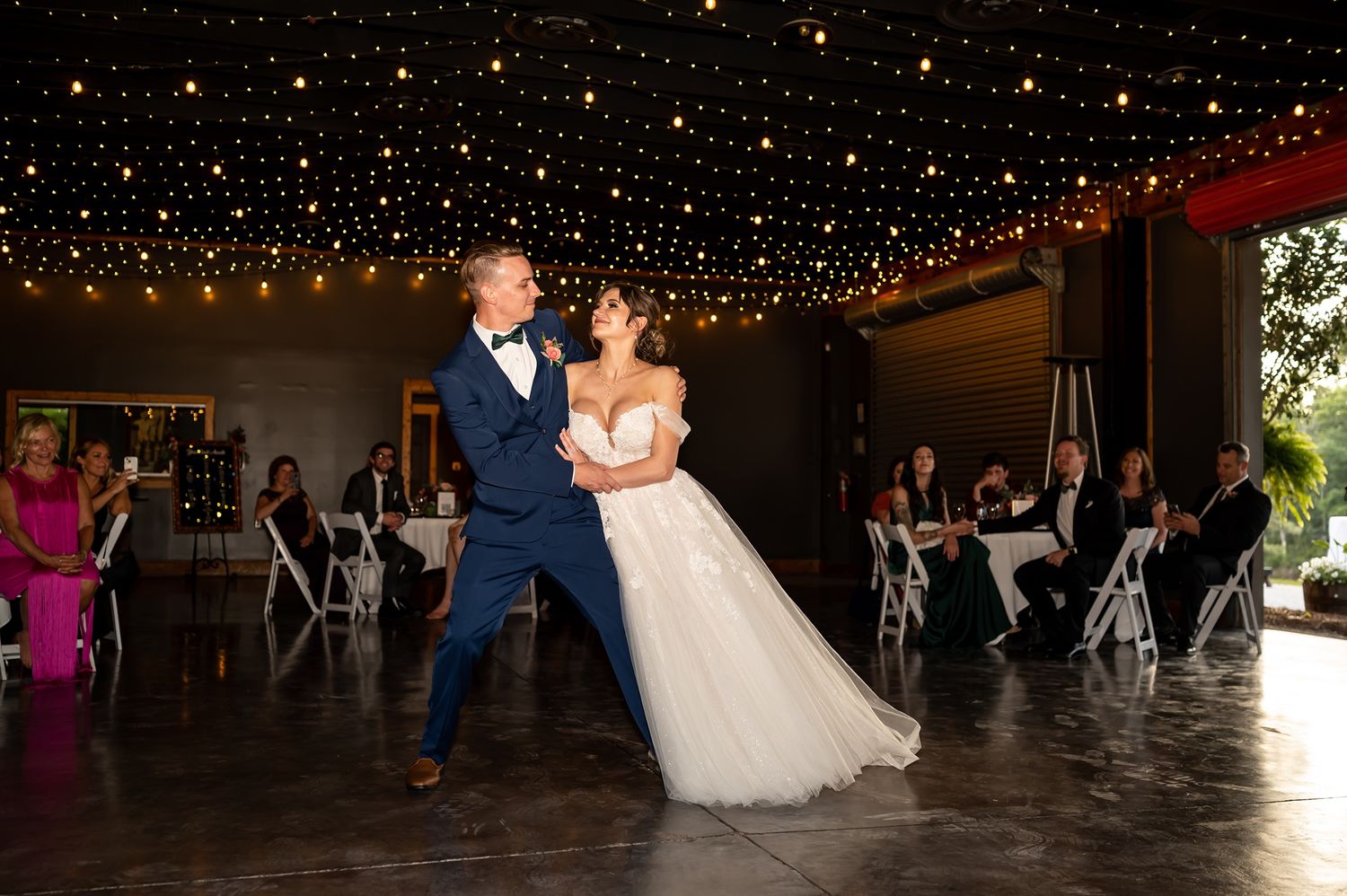 Two people dance together during their first dance at a wedding reception under twinkling string lights.