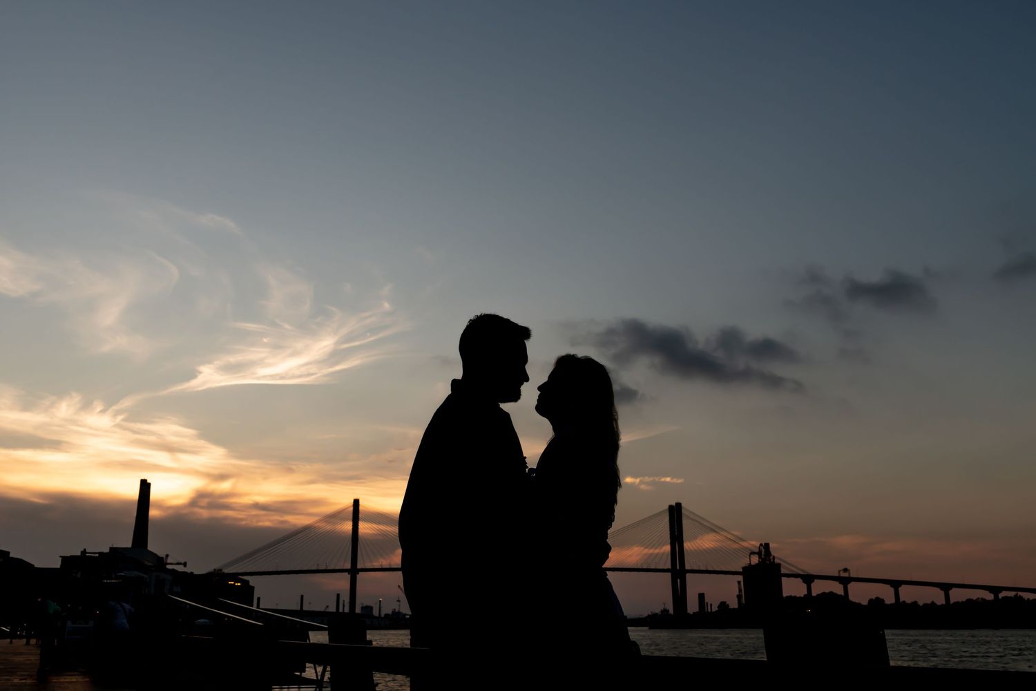 A silhouetted couple in a romantic embrace stands against a dramatic sunset sky with a bridge in the background.