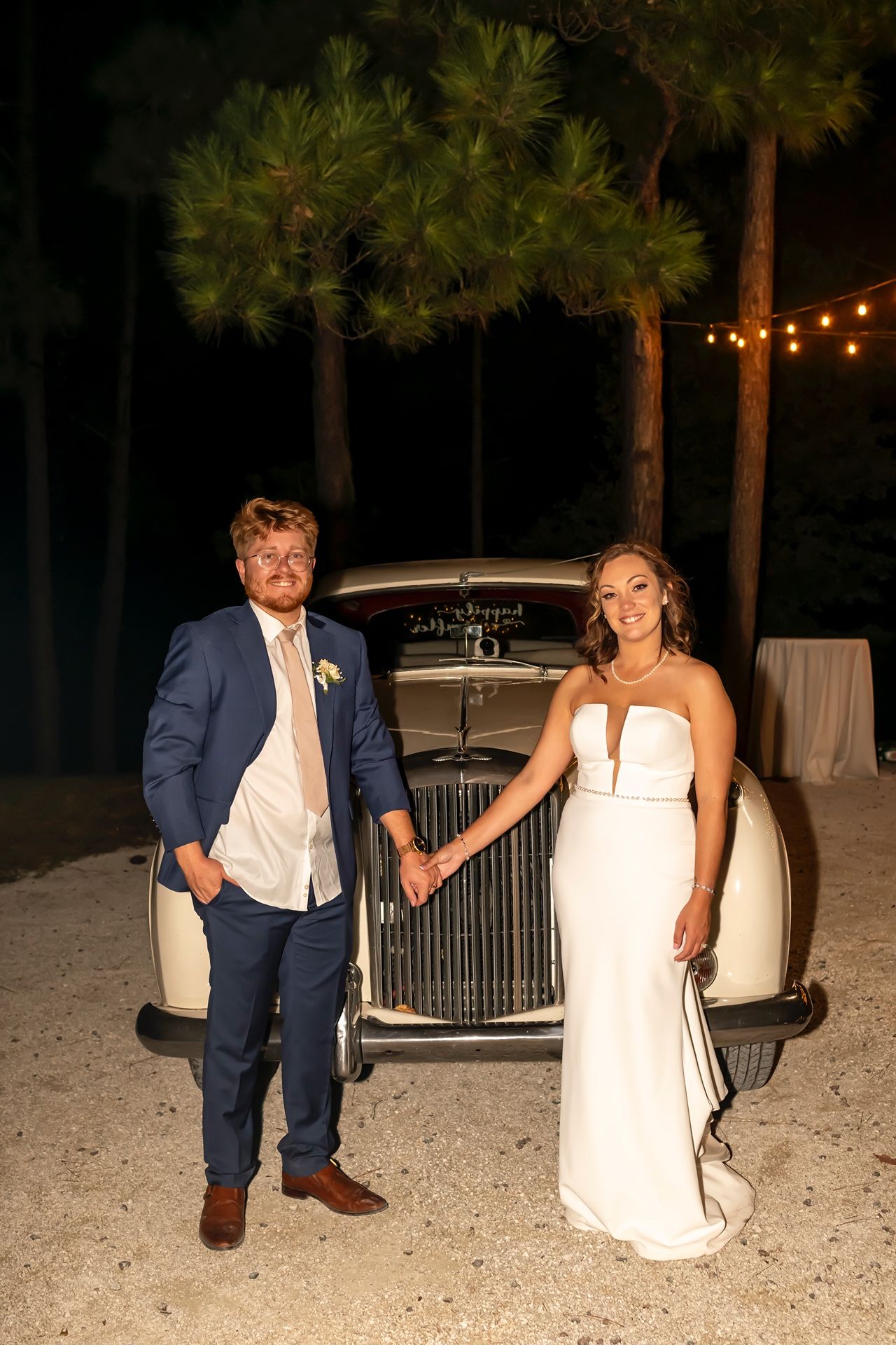 A couple in wedding attire poses together on a beach at dusk with string lights glowing in the background.