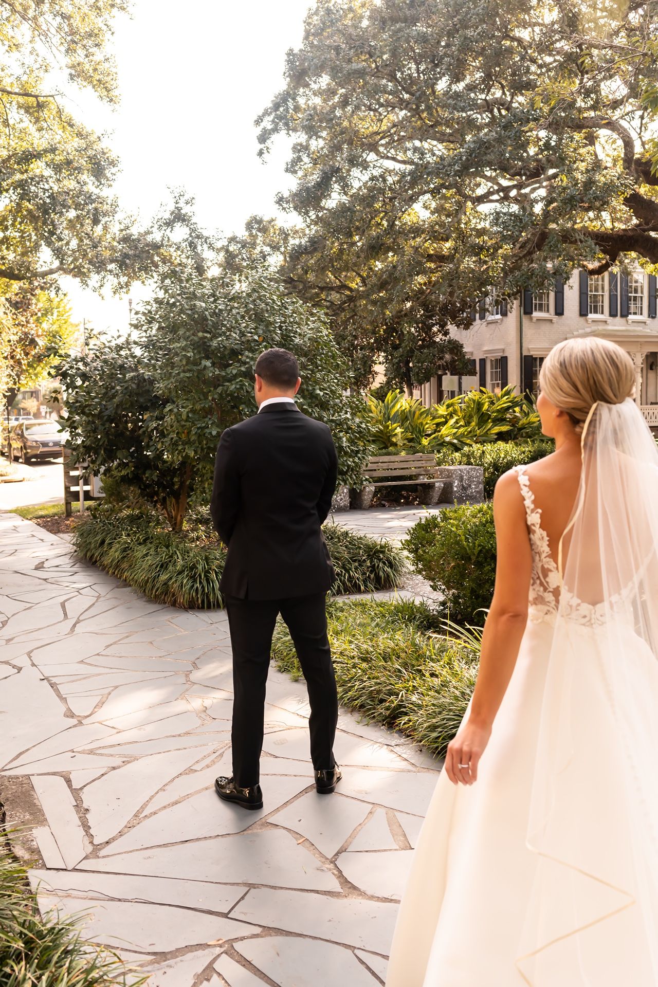 A couple in wedding attire during their first look moment on a stone pathway surrounded by lush greenery.
