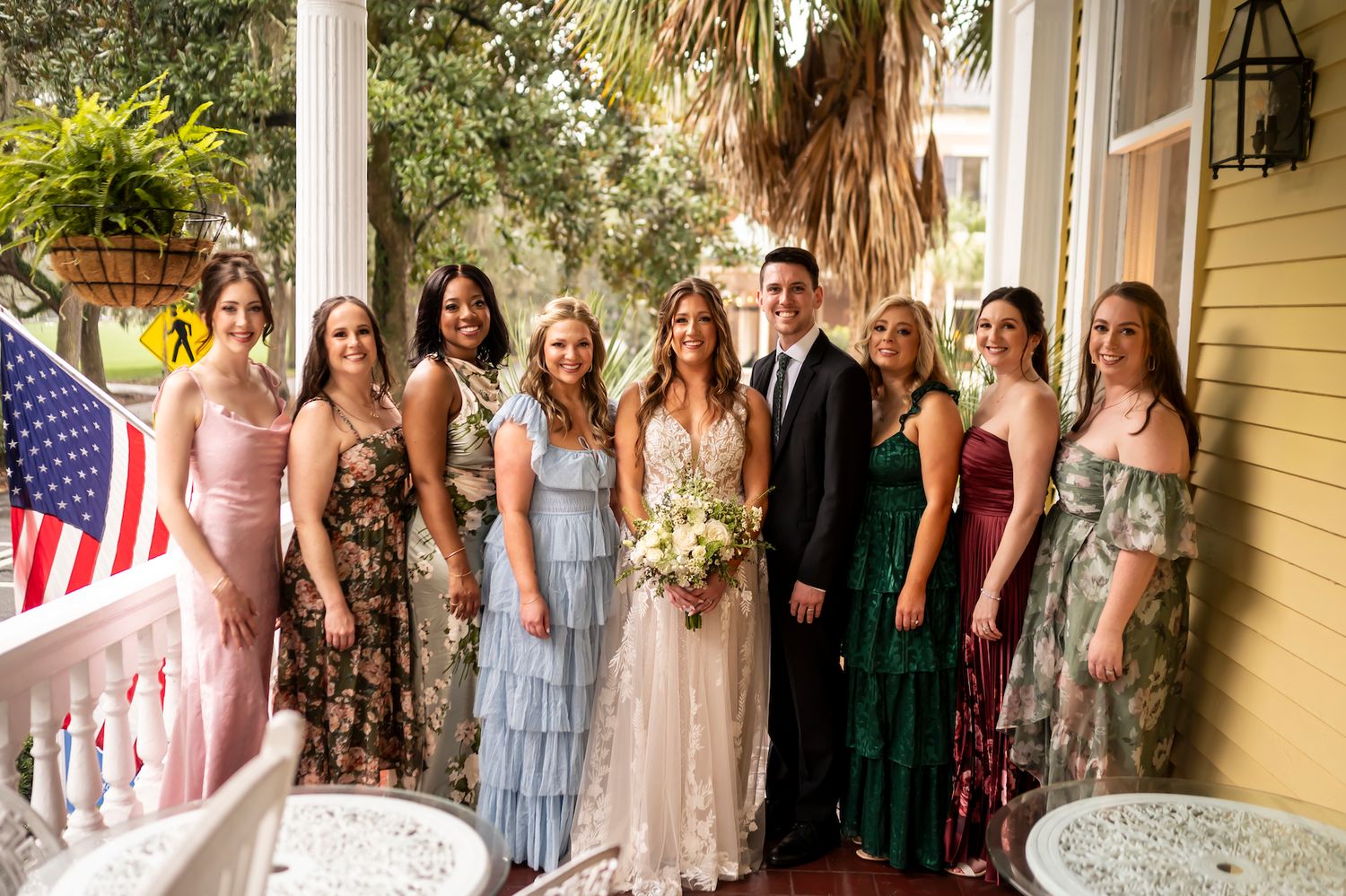 A wedding party poses together on a sunny porch, with bridesmaids wearing different colored dresses.