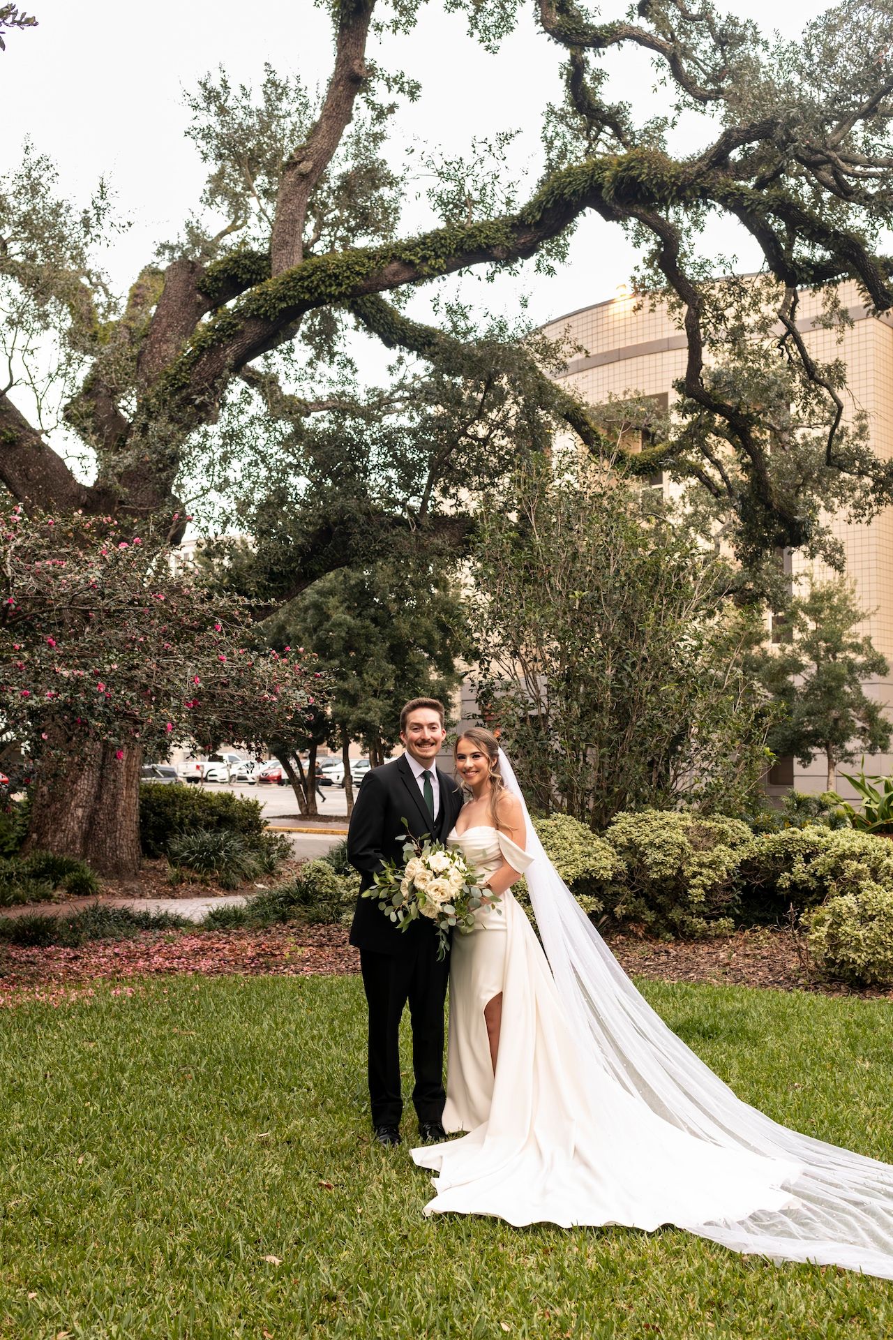 Wedding couple poses under a large oak tree on a manicured lawn with a beautiful cathedral-length veil trailing behind.