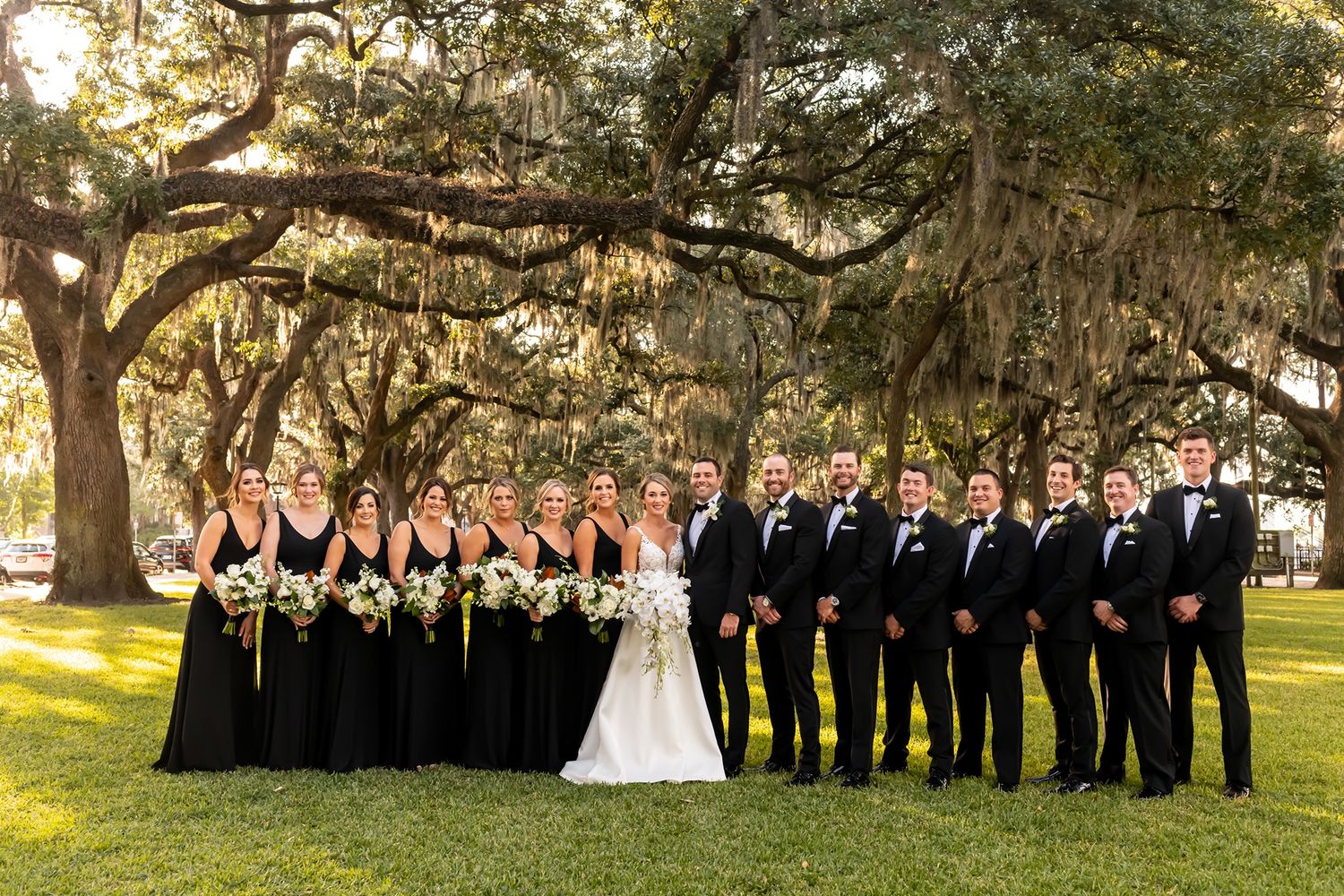 Large wedding party in black formal attire poses outdoors under Spanish moss-draped oak trees in a Southern park.