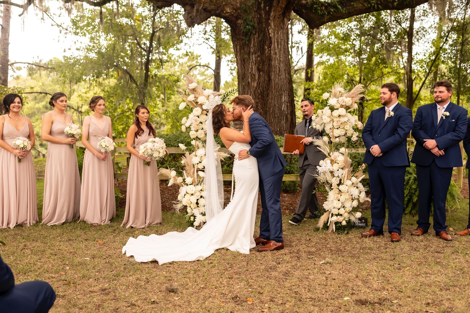 A couple embraces during their outdoor wedding ceremony under a large tree decorated with white floral arrangements.