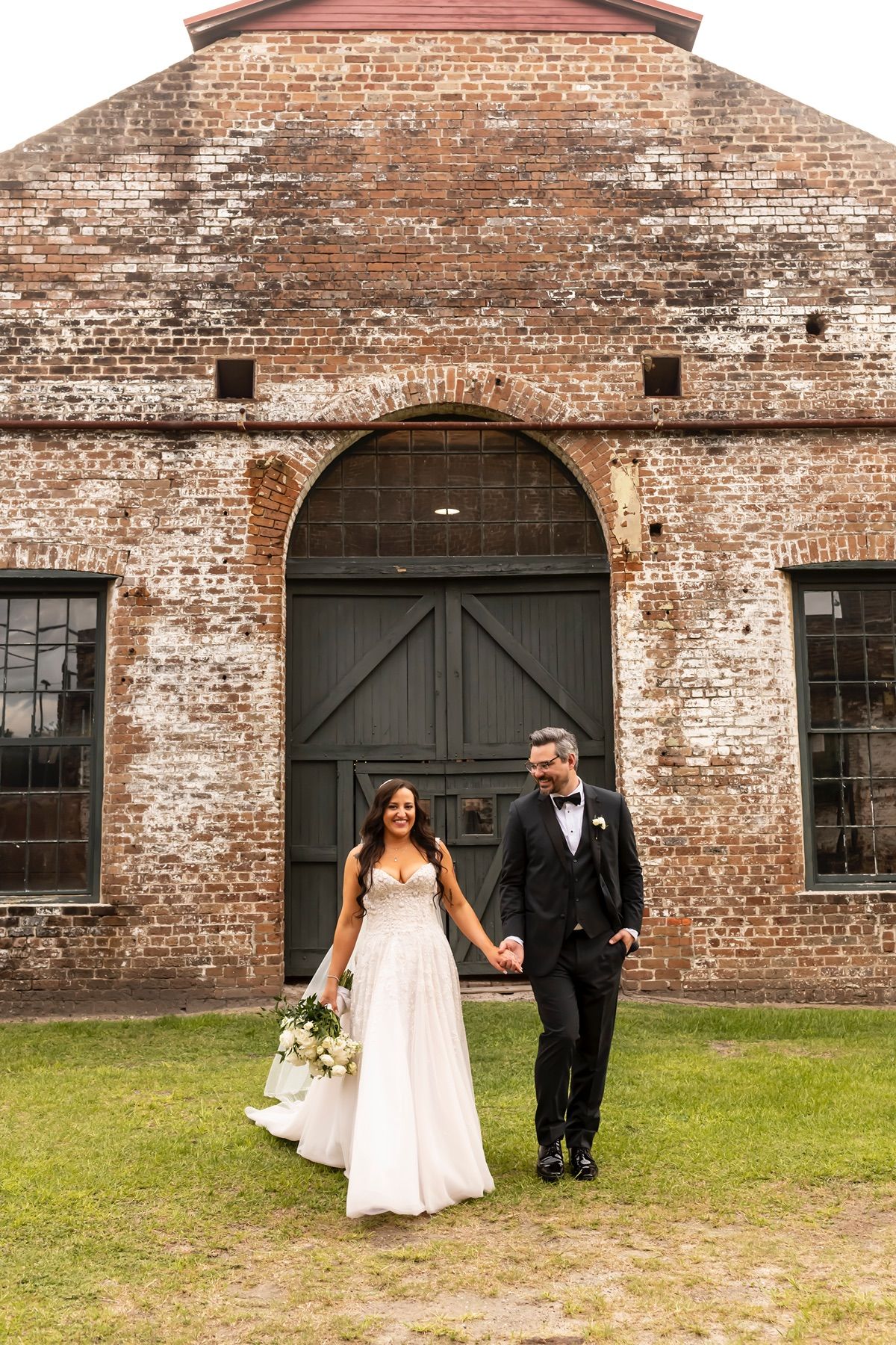 A couple in wedding attire holds hands in front of a rustic brick warehouse building with arched doorway and grassy lawn.