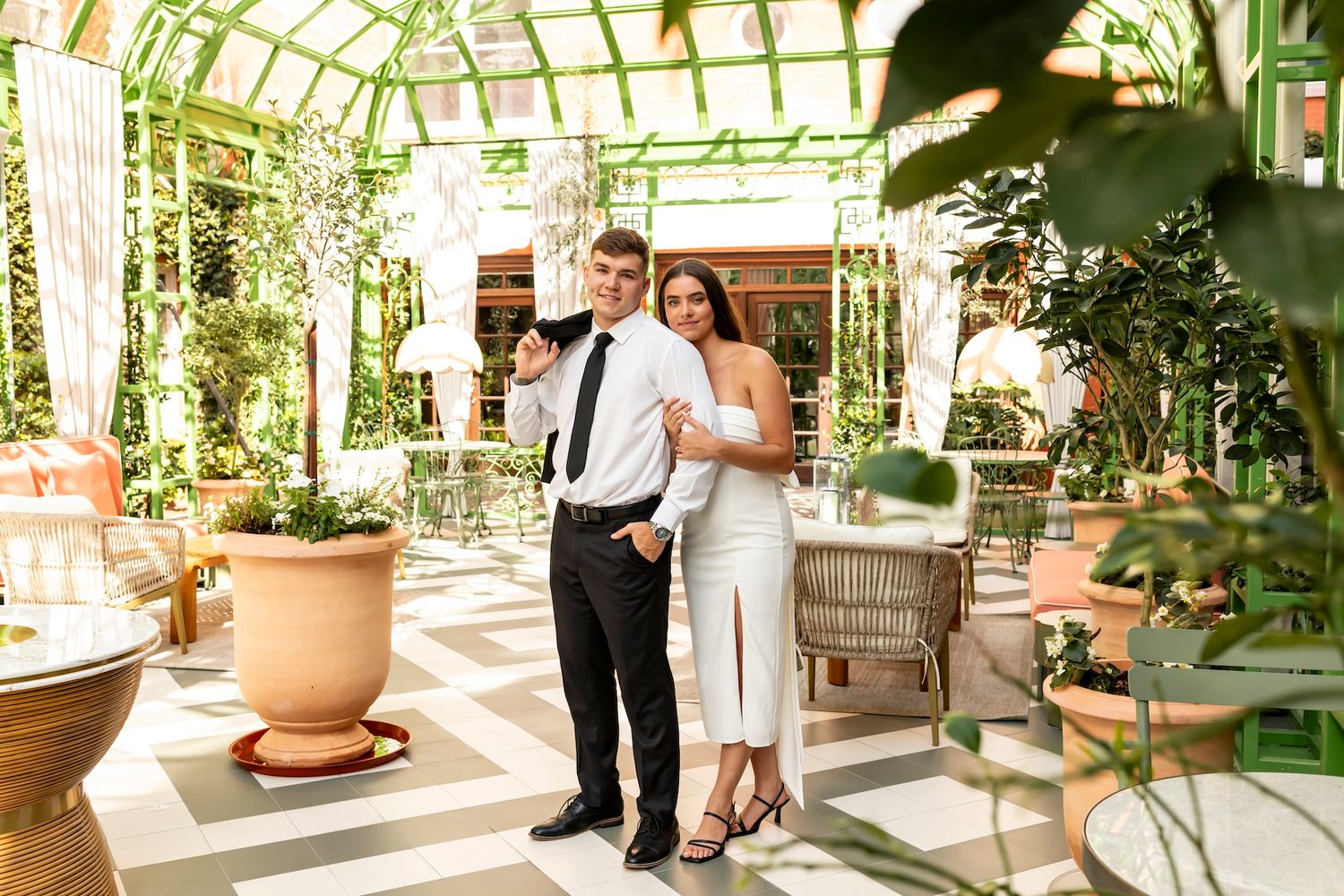 Two people in formal attire stand close together in a bright sunlit greenhouse with terracotta pots and lush plants.