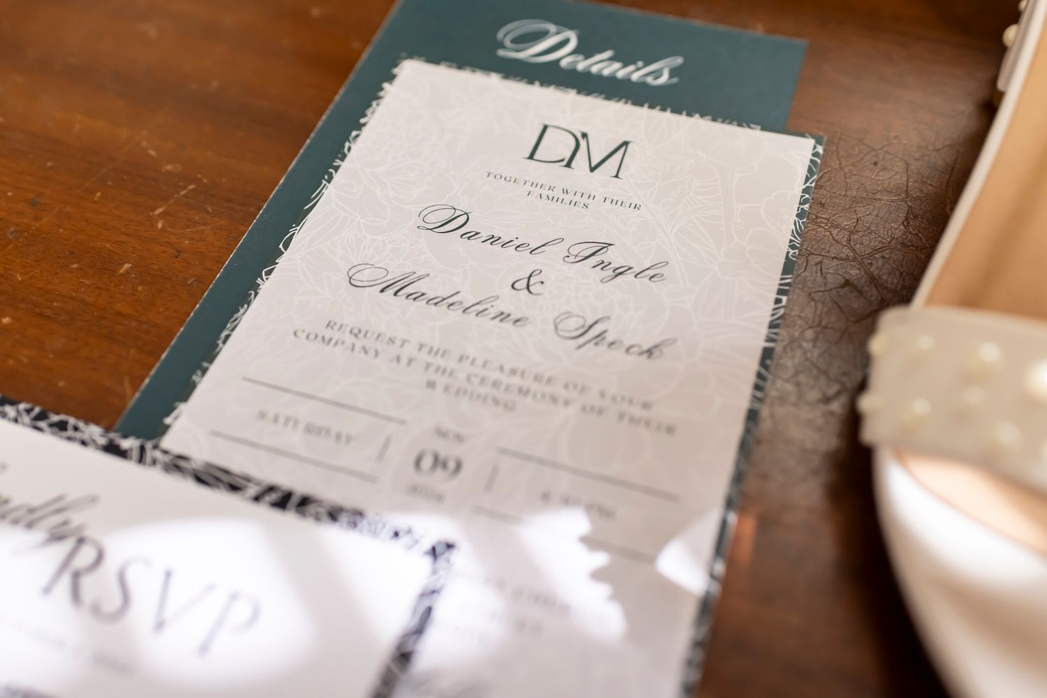 An elegant wedding invitation in teal and white sits on a wooden surface next to white fabric.