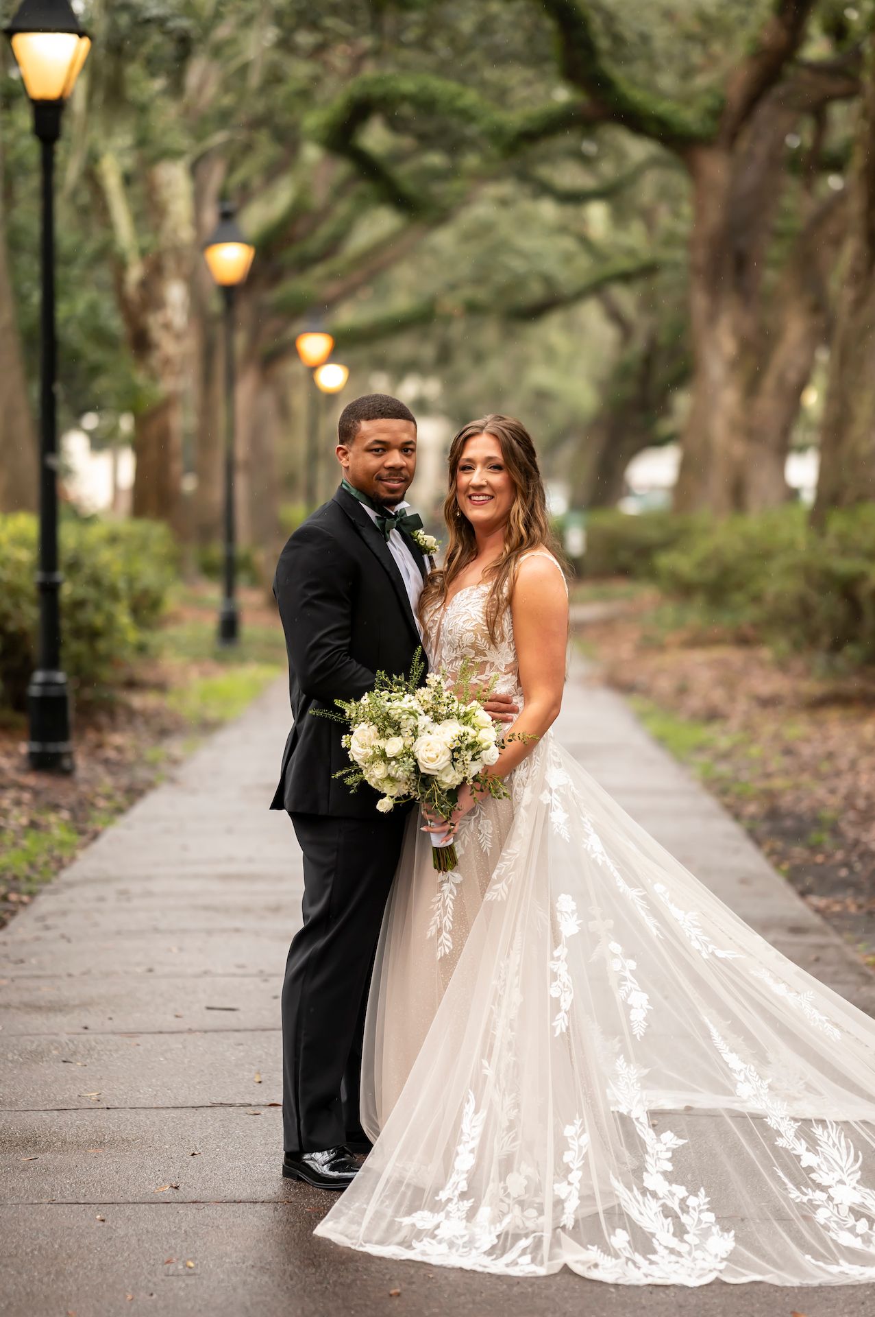 A romantic wedding couple poses on a tree-lined path with glowing lanterns at dusk.