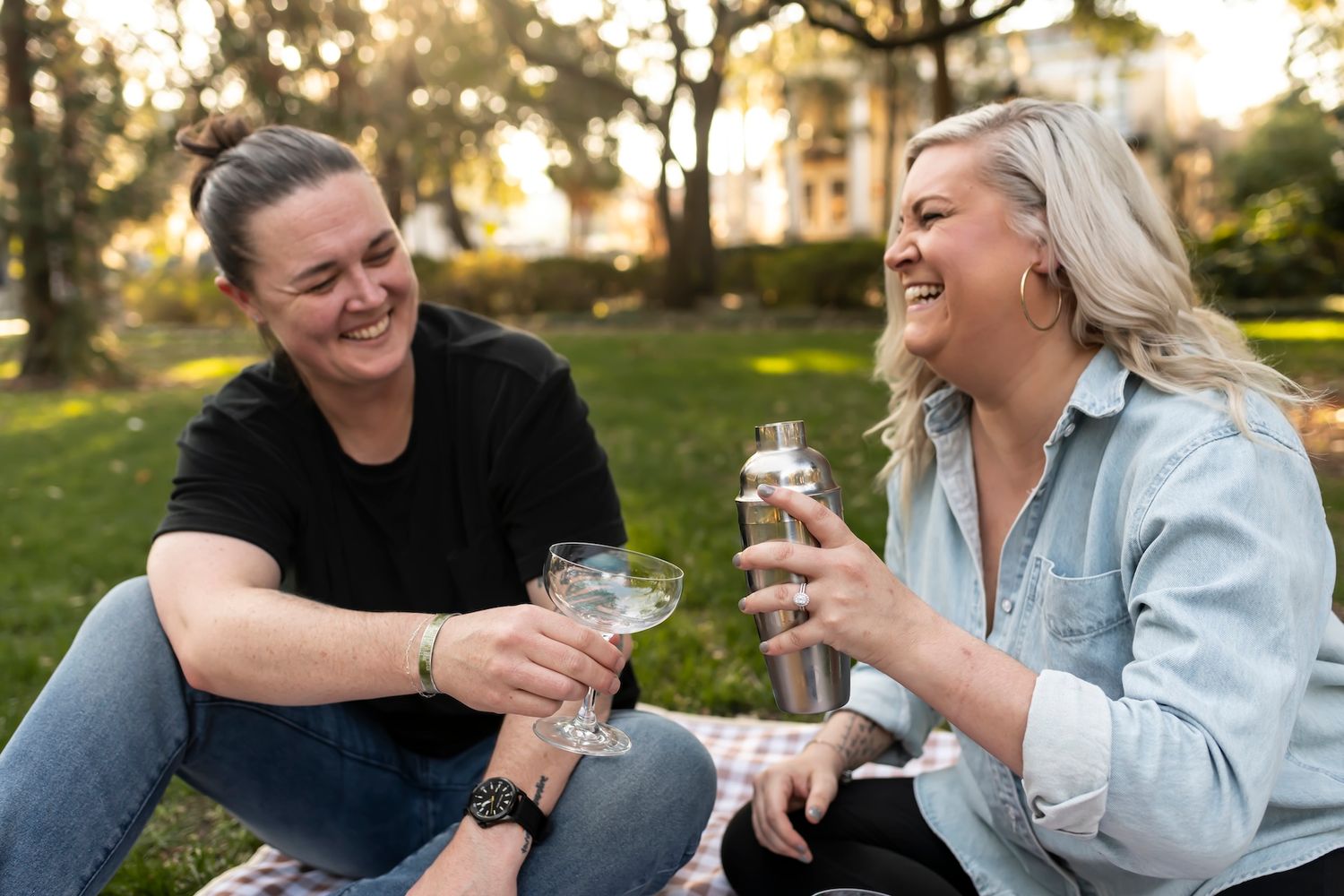Two friends enjoying wine glasses while sitting on grass during golden hour sunset in a park.