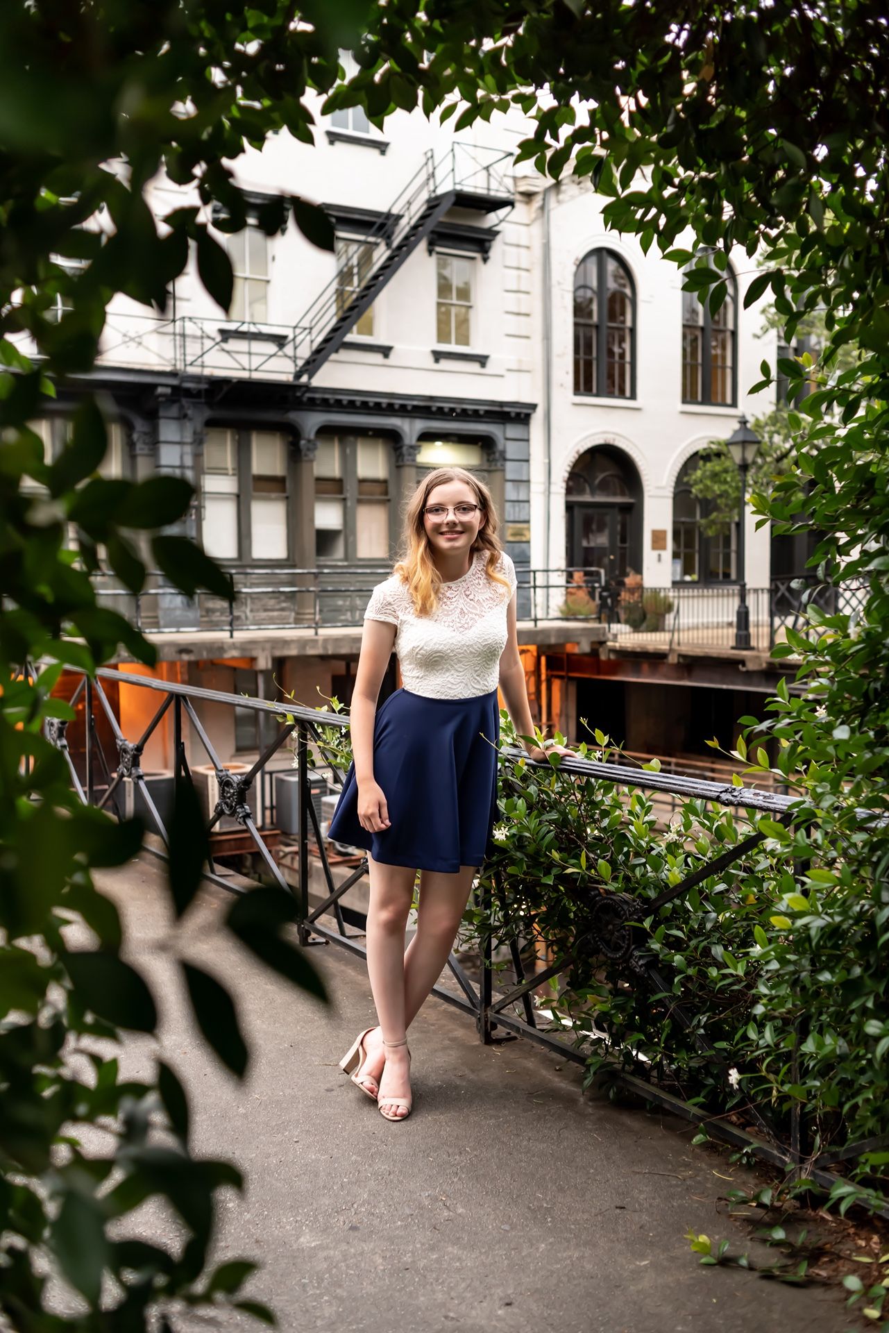 A woman in a white blouse and navy skirt smiles while standing on a bridge near classic architecture.