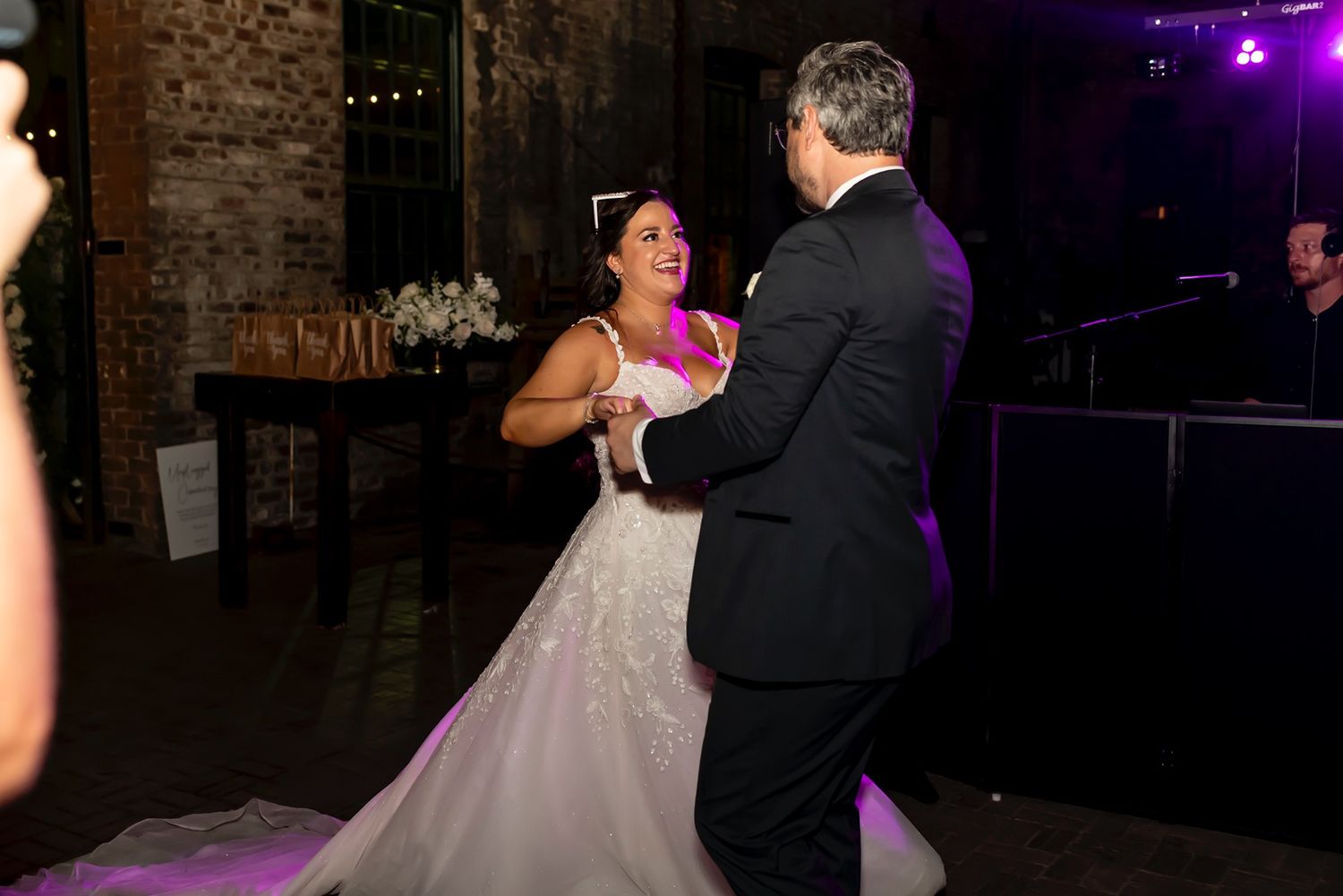 A couple shares a joyful moment dancing together in a rustic brick-walled venue with purple lighting.