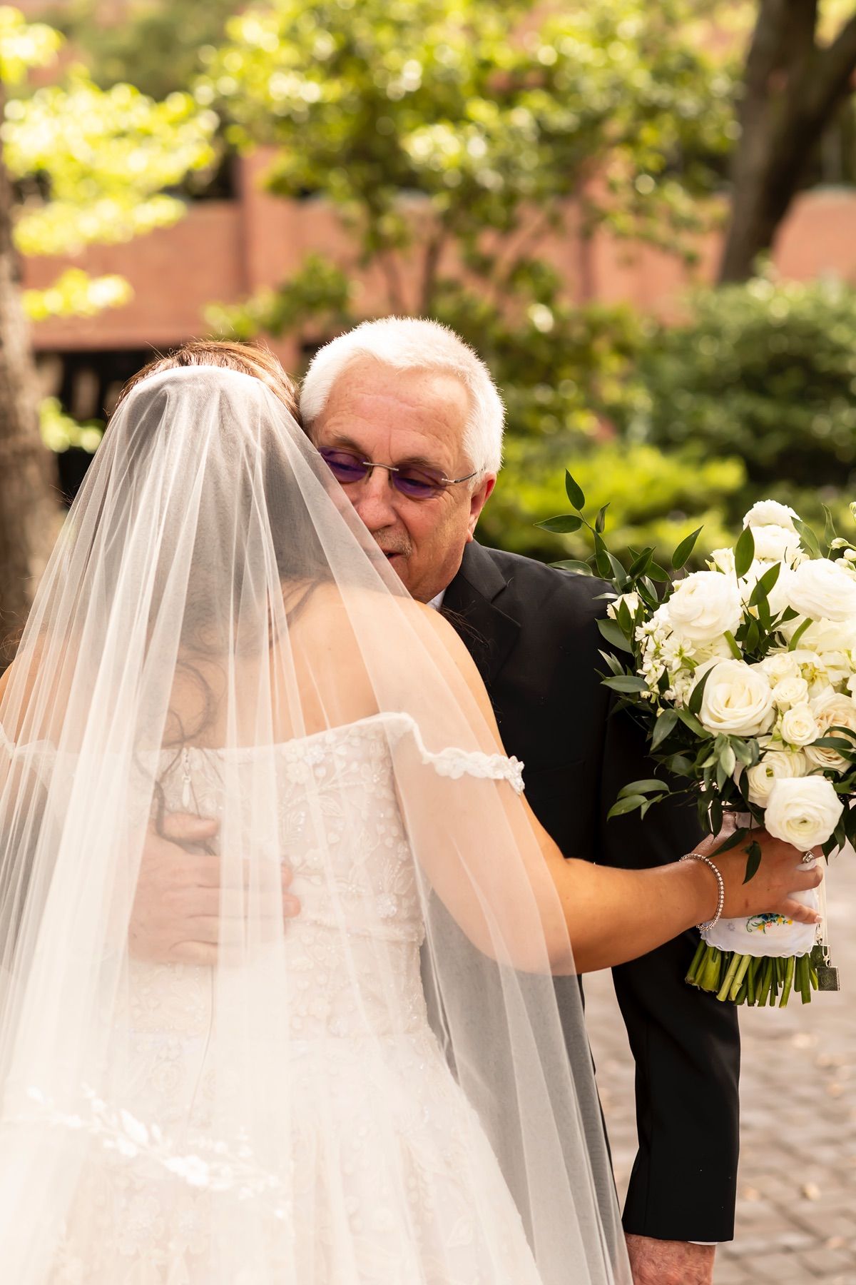 A person in a white wedding dress holding a bouquet embraces someone in a dark suit during an outdoor ceremony.