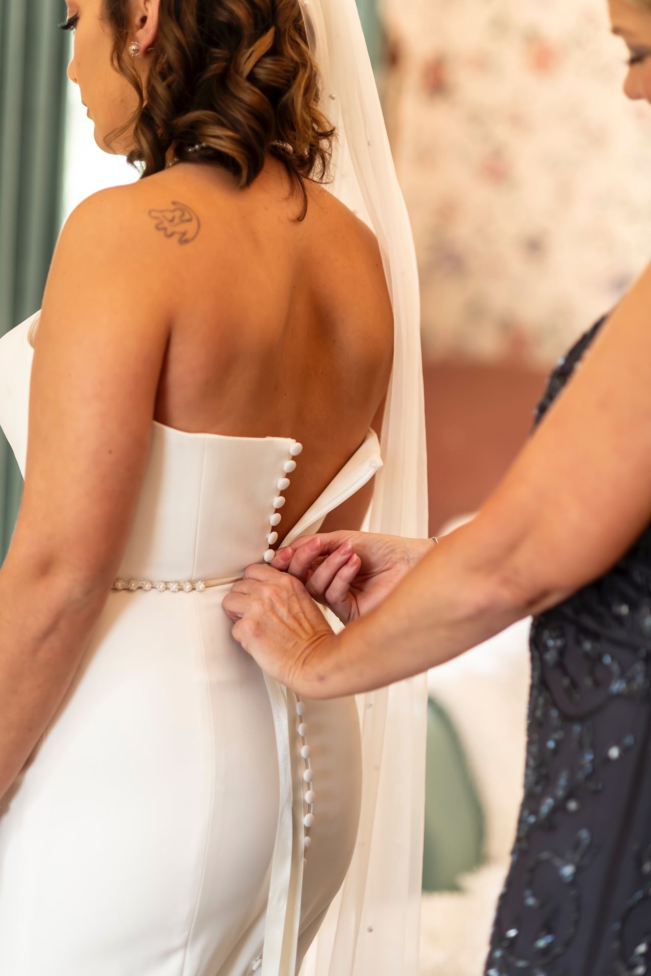 Someone helps button up an elegant white strapless wedding dress with delicate details on a bride's back.