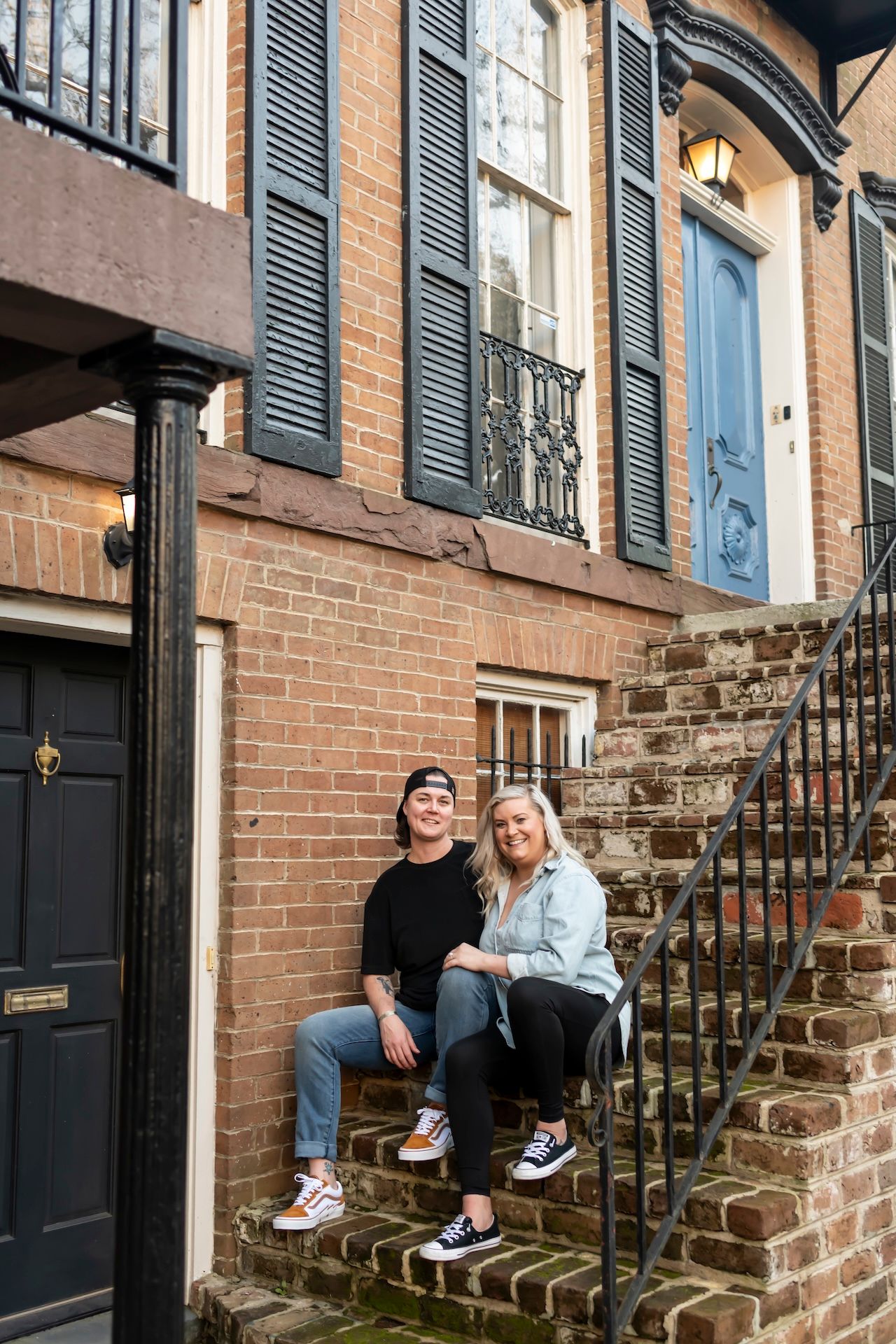 Two people sit casually on historic brick steps outside a charming row house with blue shutters in an urban setting.